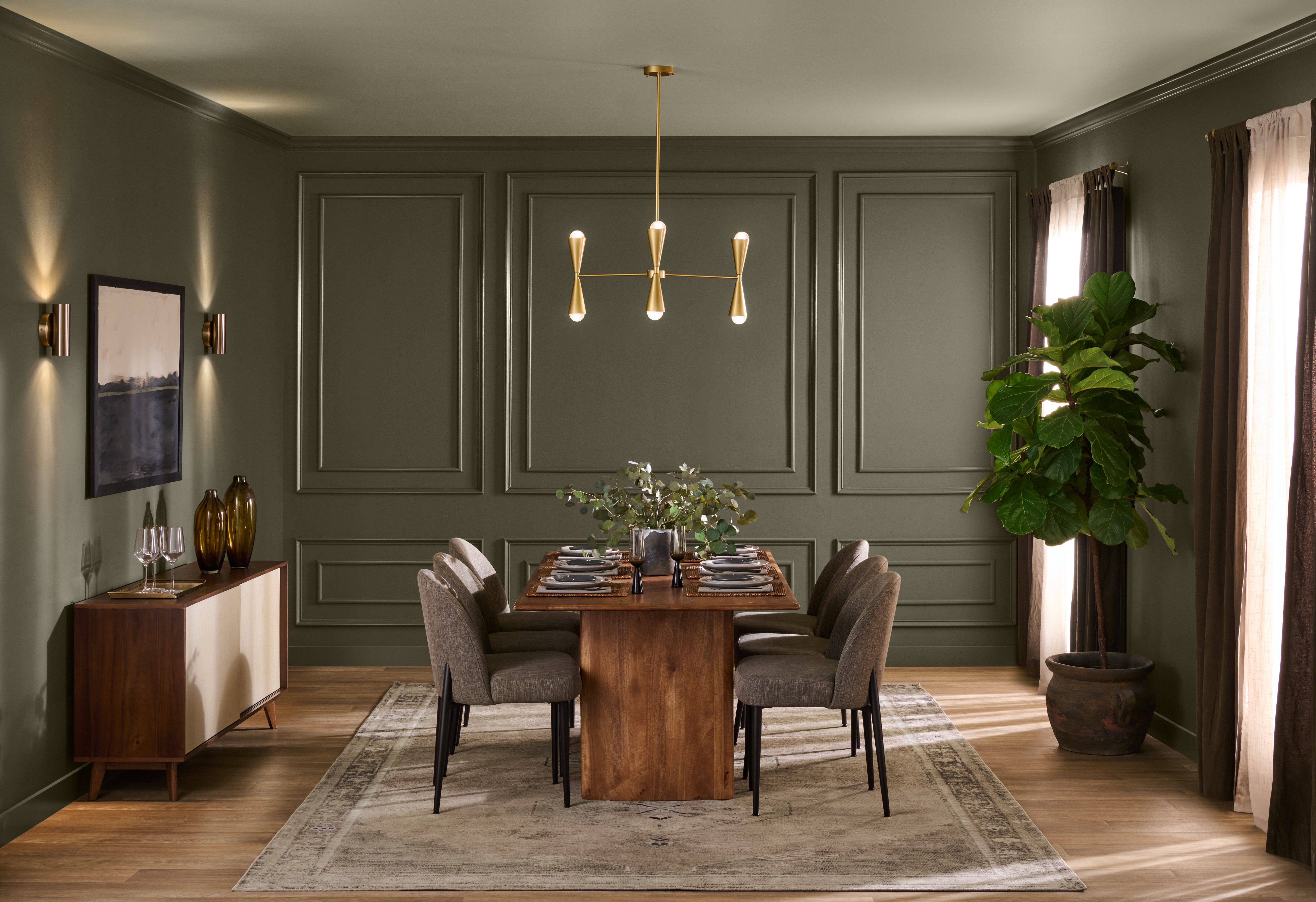 Cette image montre une élégante salle à manger baignée d’une lumière naturelle chaleureuse avec des murs lambrissés peints en teinte Terre à terre de BEHR, ainsi qu’une table à dîner en bois massif entourée de chaises rembourrées et surplombée d’un lustre moderne en laiton.
