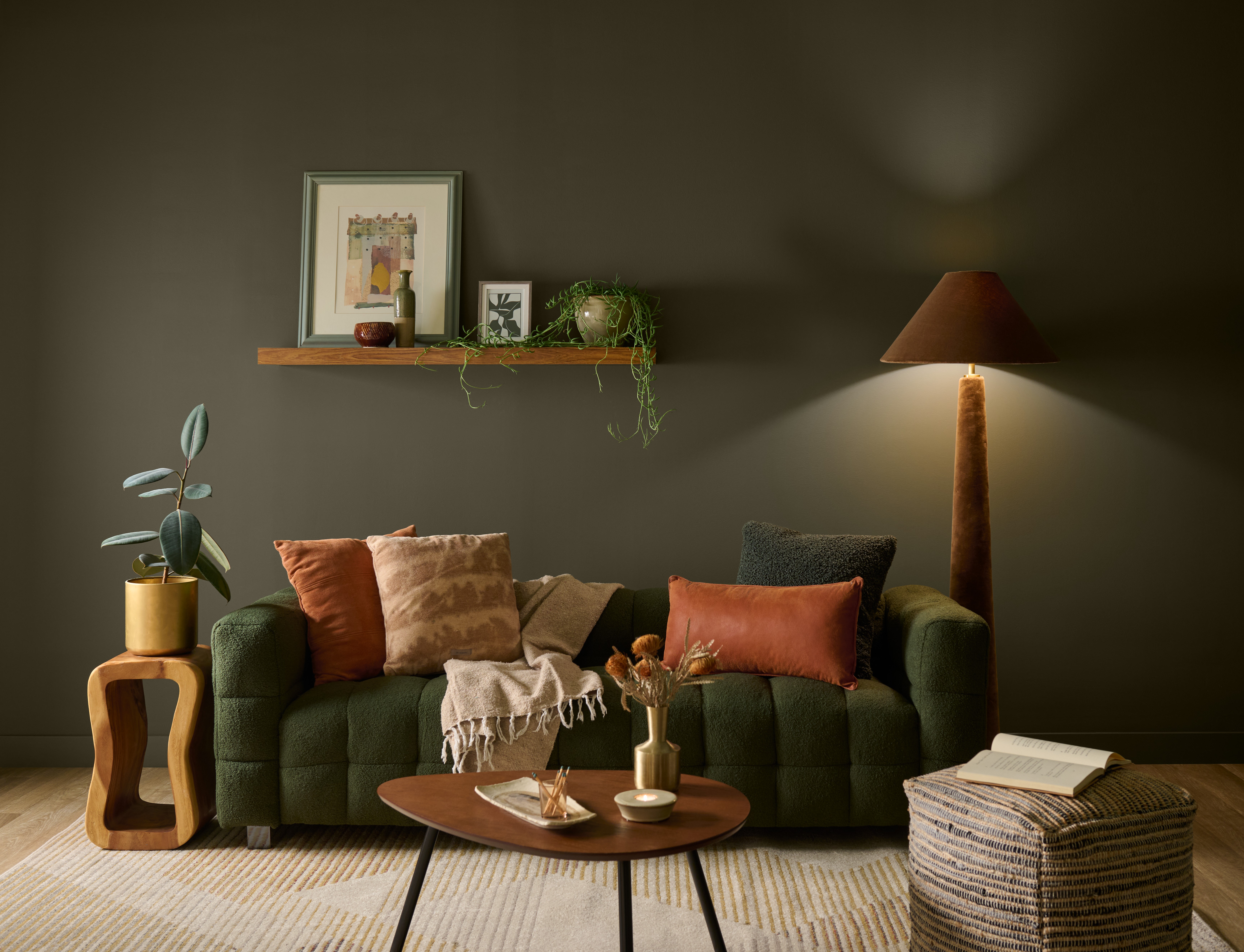 Cette image montre un salon douillet dont le mur peint en teinte Terre à terre de BEHR s’assortit d’un sofa vert foncé, de coussins rouille au ton chaud, d'une table basse en bois, d’un amalgame de textures naturelles et d’un éclairage d’ambiance focalisé.