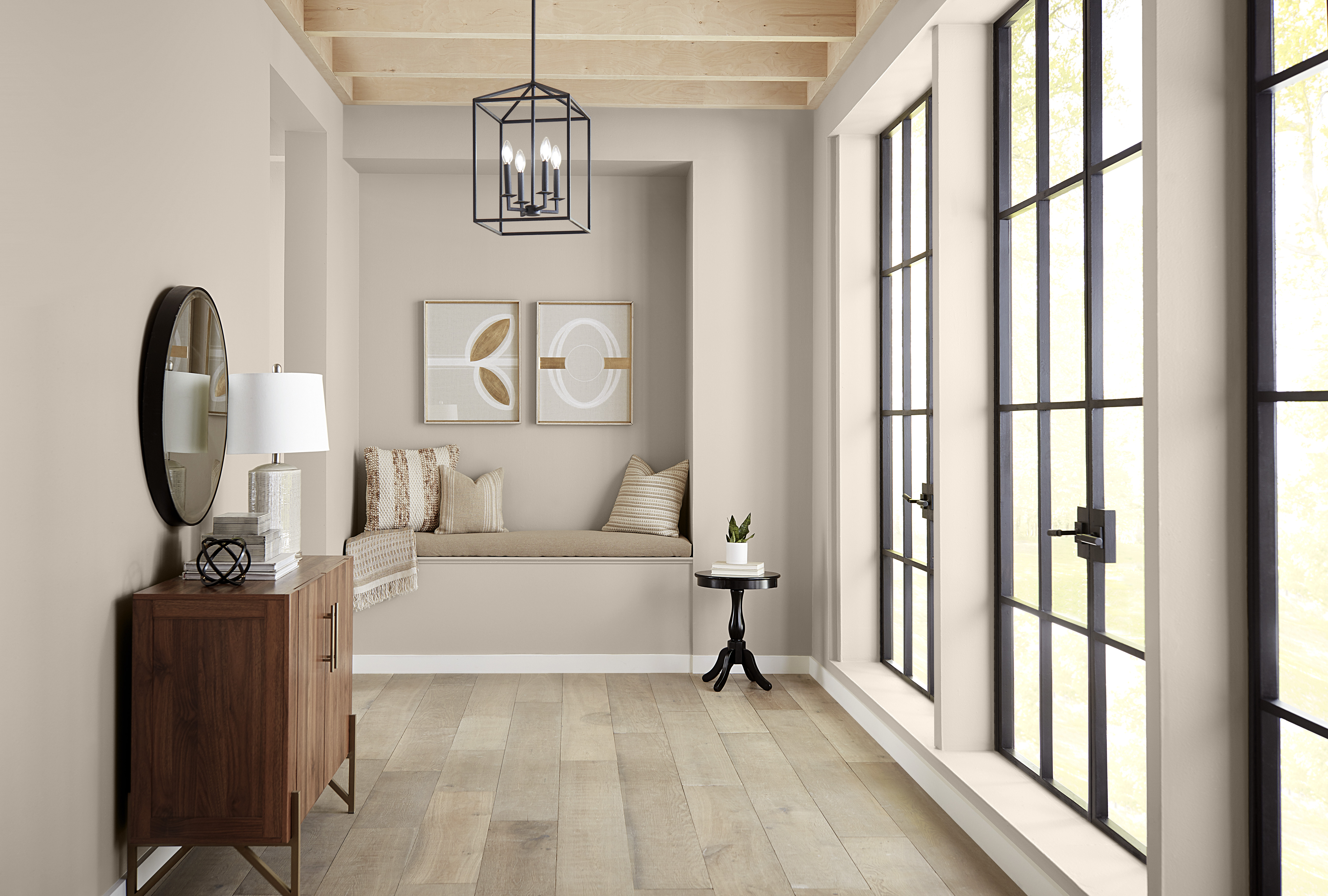 Cette image montre un couloir baigné de lumière dont les murs peints en Pain de blé créent une toile de fond neutre et chaleureuse qui met en valeur les fenêtres à encadrement noir, le parquet de bois clair, les solives apparentes au plafond et un coin boudoir en alcôve serti de coussins confortables aux teintes neutres.