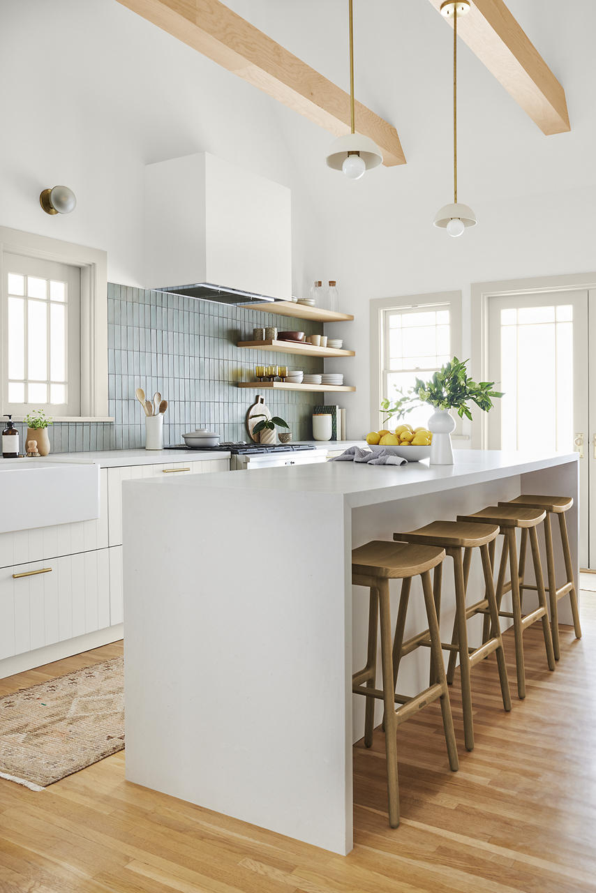 Cette image montre une cuisine de style «ferme bio moderne» au décor hautement texturé et nuancé, avec des murs blancs, des armoires blanches, un grand îlot gris pâle, des poutres apparentes de couleur claire, des tablettes en bois, des tabourets de bar en bois, un dosseret bleu pâle et un parquet en bois naturel au ton chaud.