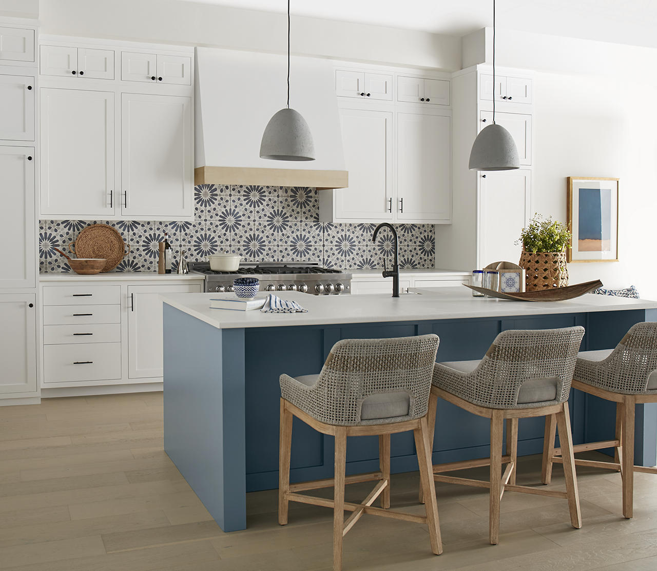 Cette image montre une cuisine à tonalité blanche avec un îlot peint en une populaire teinte de bleu cendré, un dosseret en céramique à motif bleu et blanc, des armoires blanches, deux luminaires suspendus gris, trois chaises de bar en rotin et un parquet en bois blond.