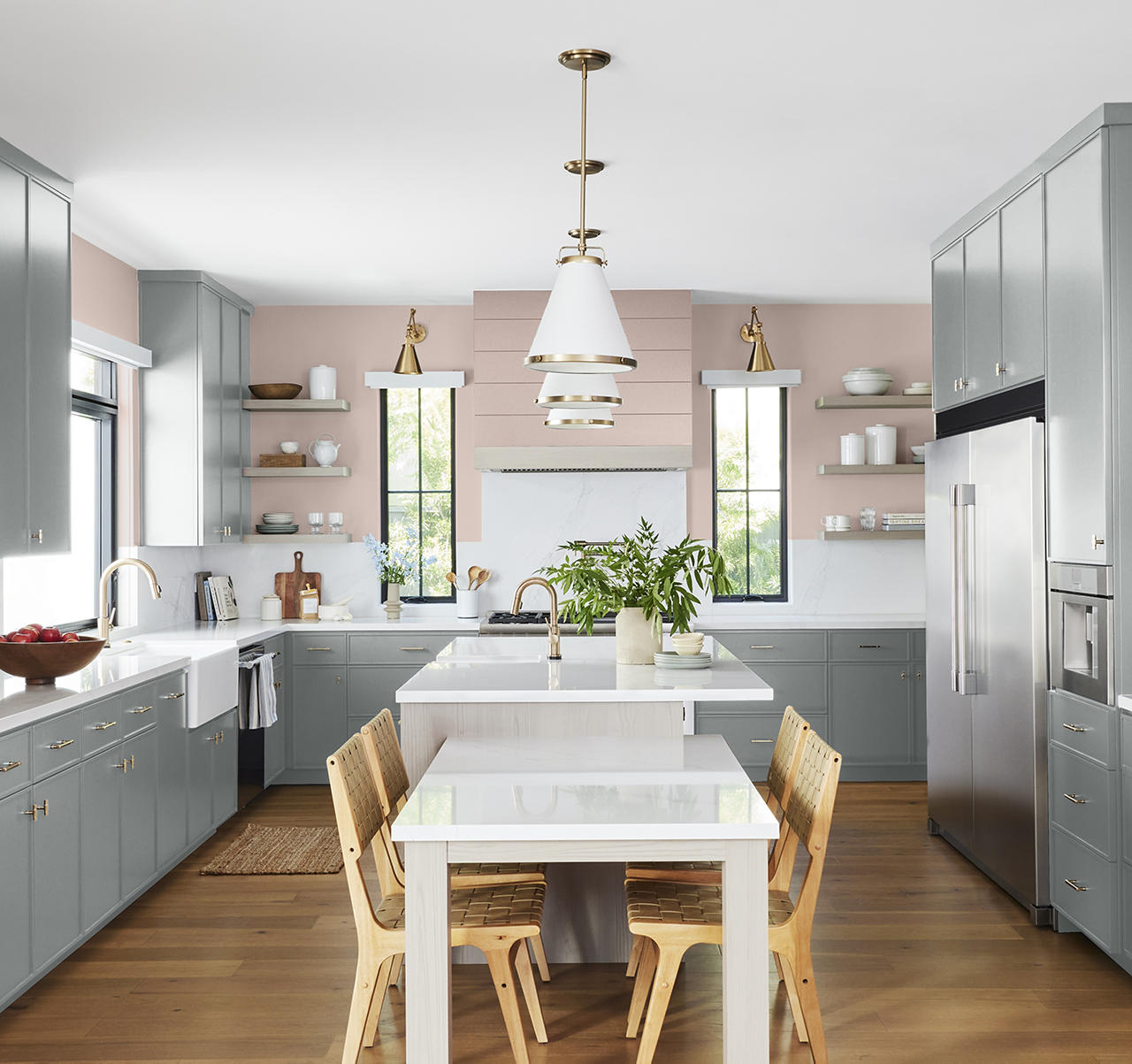 Cette image montre une cuisine lumineuse avec des armoires gris-bleu, des poignées en laiton, des comptoirs blancs, un mur d’accent rose pâle, des chaises de salle à manger en bois et une abondante lumière naturelle. Les armoires gris-bleu ancrent l’espace dans une ambiance apaisante et sophistiquée, tandis que le mur rose pâle apporte une douceur candide, le tout formant un décor équilibré qui inspire un sentiment de fraîcheur et de convivialité. C’est l’agencement de couleurs idéal pour ajouter de la profondeur et du caractère à cette cuisine.