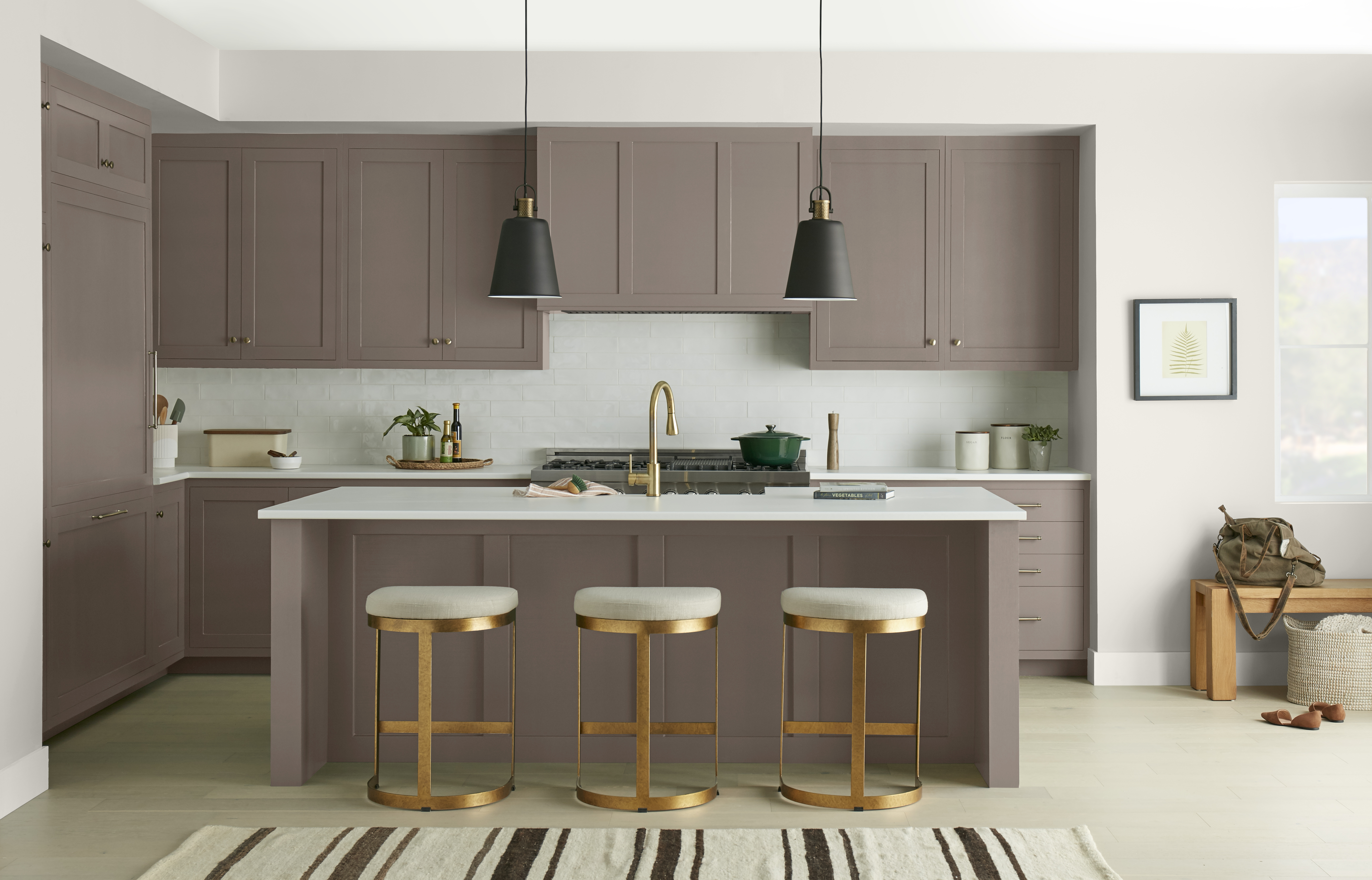 Cette image montre une élégante cuisine peinte en brun taupe , avec des armoires allant du sol jusqu’au plafond, un îlot de même couleur, des comptoirs blancs, des accessoires en laiton et deux luminaires noirs suspendus au-dessus de l’îlot. L’amalgame de ces tons neutres crée une ambiance raffinée et sophistiquée.