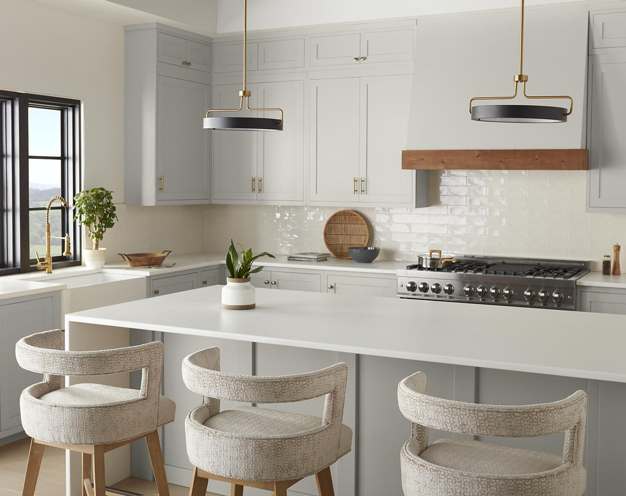 Cette image montre une cuisine aux tons de blanc et de gris clair, agrémentée d’une moulure en bois naturel au-dessus de la cuisinière, de poignées en laiton, de luminaires suspendus en laiton, de comptoirs blancs et de chaises de bar de couleur neutre. Les nuances du bois tranchent contre la monochromie des armoires grises, tandis que les accessoires en laiton apportent une touche moderne et raffinée à l’ensemble.