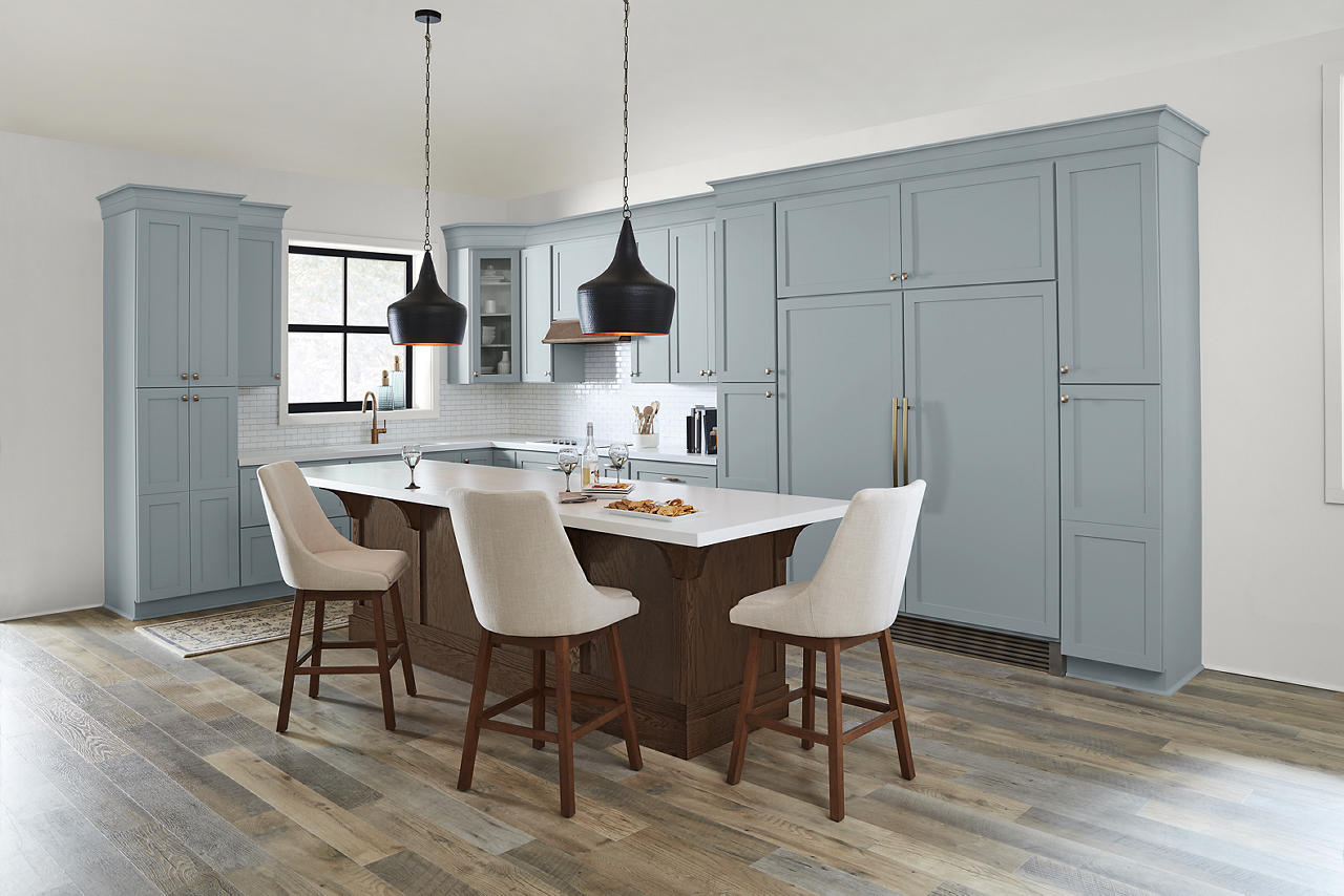 Cette image montre une cuisine avec des armoires peintes en une populaire nuance de bleu pâle et munies de poignées en laiton brossé, ainsi qu’un îlot en bois sombre, un parquet aux tons chauds, deux luminaires suspendus noirs et trois chaises de bar de couleur crème.