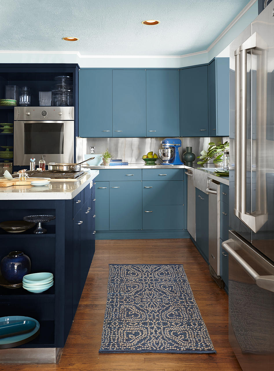 Cette image montre une cuisine inondée de bleu avec un îlot dont les armoires sont peintes en une riche nuance de bleu marine. Les armoires fixées au mur sont peintes en un bleu plus pâle et le plafond est peint en un subtil ton de bleu crème, le tout créant un décor audacieux et immersif. Un tapis d’appoint à motif bleu et blanc ainsi qu’une vaisselle bleue viennent compléter la trame de couleurs, tandis que les électroménagers en inox et le parquet au ton chaud apportent de l’équilibre et du contraste à l’ensemble.