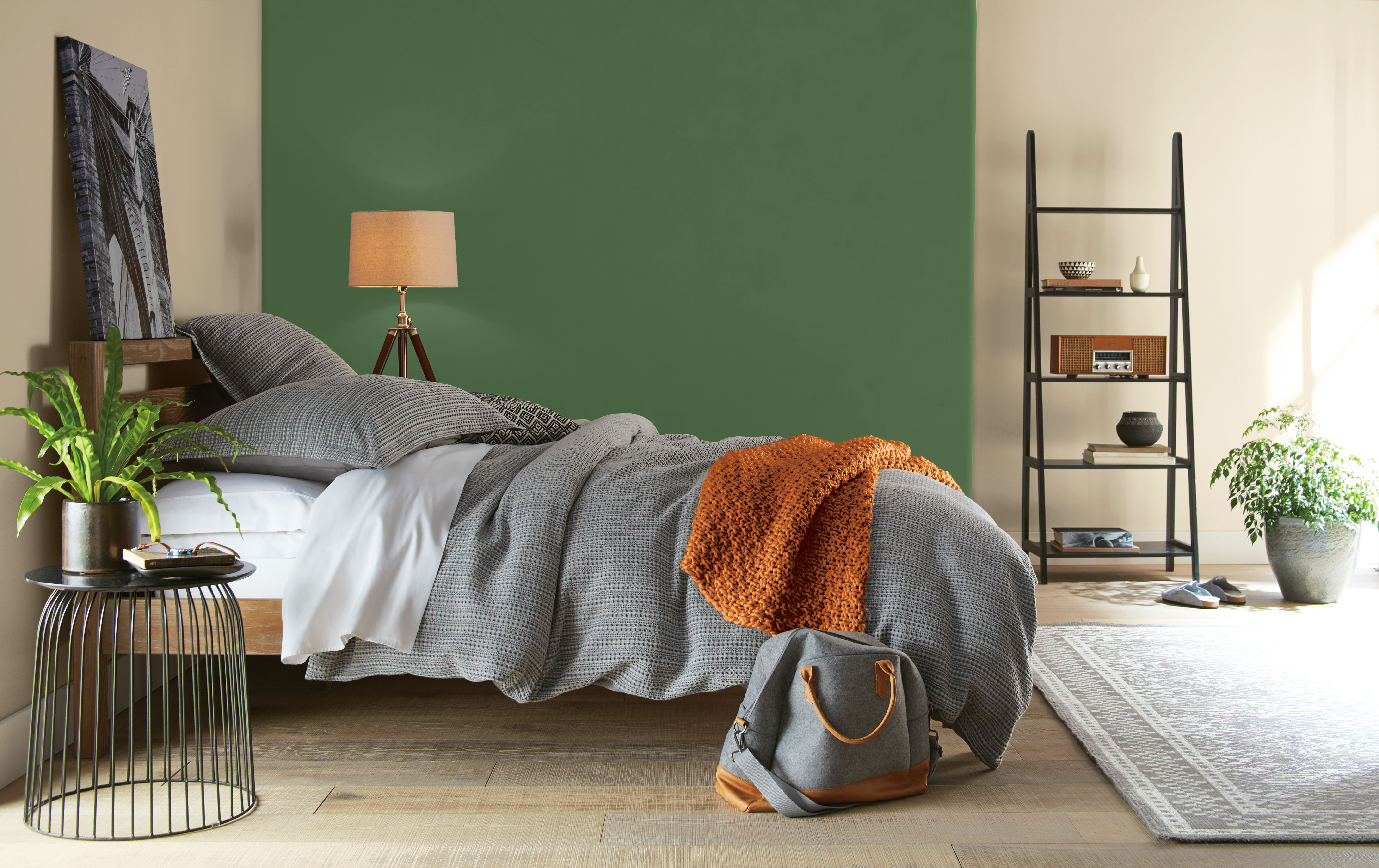 Cette image montre une chambre à coucher avec un mur de fond peint en Concombre de jardin, un vert profond qui s’assortit à merveille avec les tons neutres et chaleureux des autres murs et du plancher, la literie grise, les meubles en bois naturel, les plantes en pot et le jeté de couleur ocre orangée.