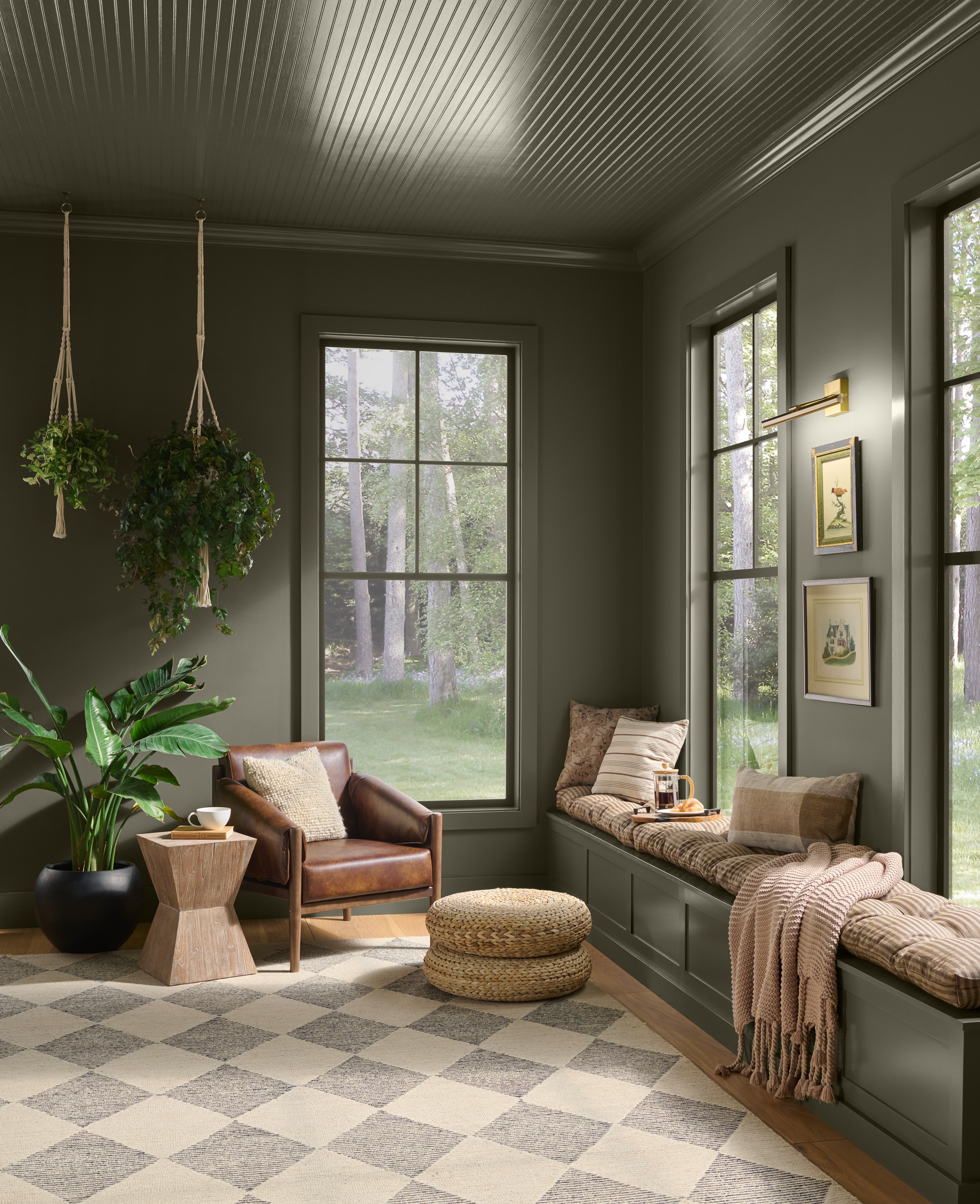 Cette image montre une véranda chaleureuse peinte en teinte Terre à terre de BEHR, une banquette encastrée sous deux grandes fenêtres, un fauteuil en cuir, des jardinières suspendues et un tapis à losanges gris et crème.
