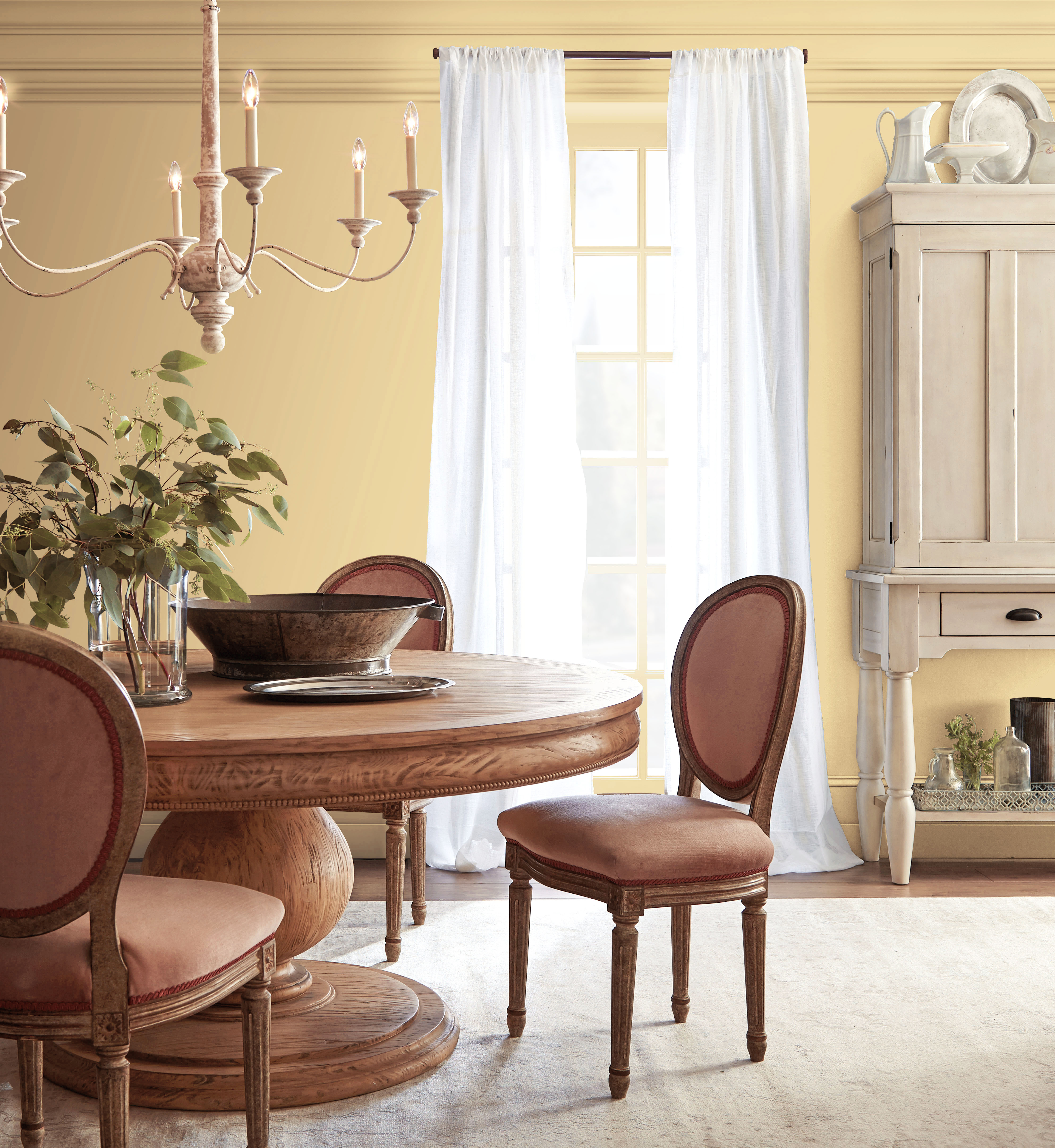 Cette image montre une salle à manger avec un mur peint en Or harmonieux, un mobilier en bois au ton chaud, des rideaux blancs et un luminaire de plafond de style chandelier, le tout ancré dans un décor romantique inspiré de l’époque de la Régence.