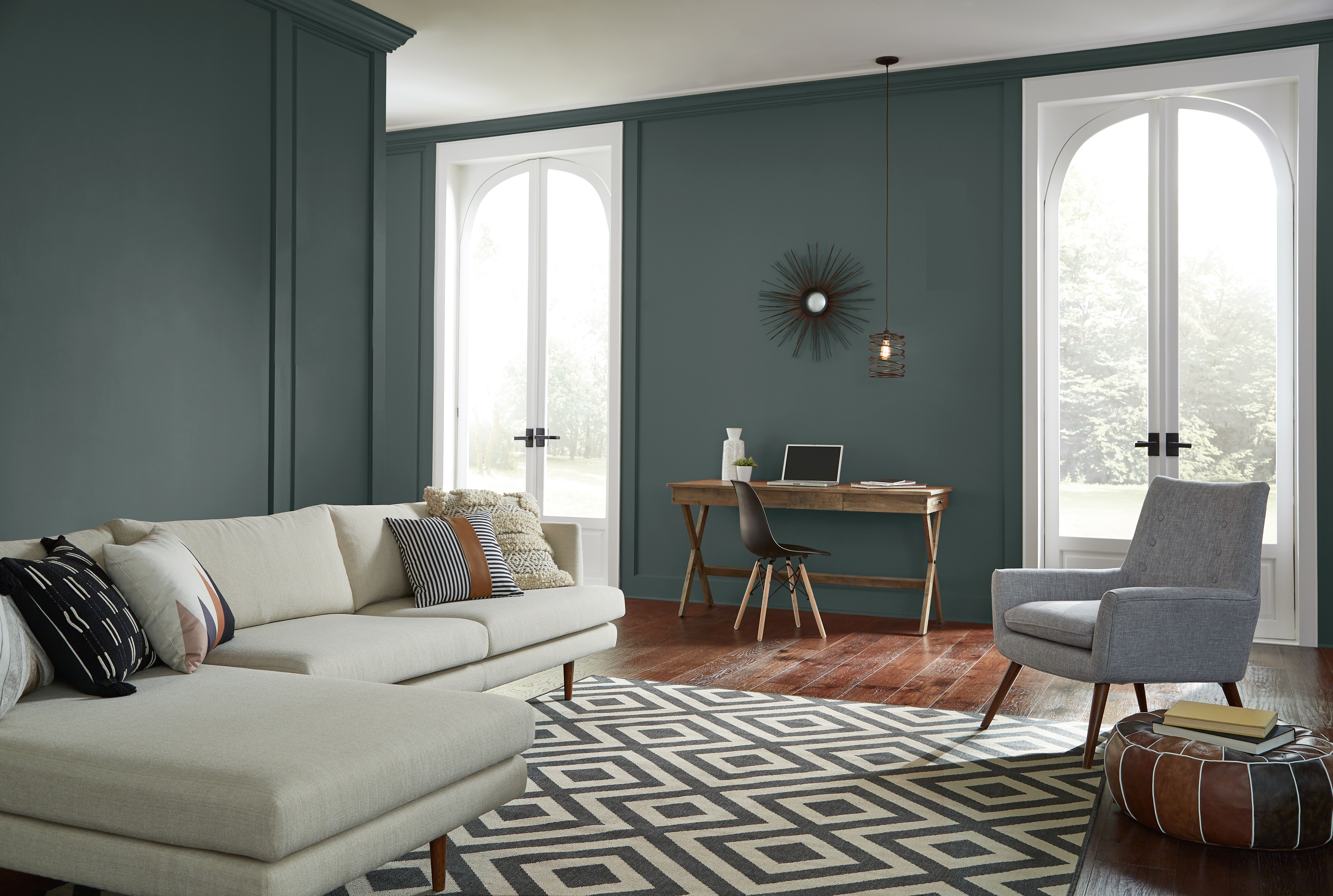 Cette image montre un grand salon ouvert avec des murs peints en vert foncé, une petite table de bureau adossée au mur entre deux grandes portes-fenêtres, un canapé sectionnel blanc, un fauteuil gris et un parquet en bois.
