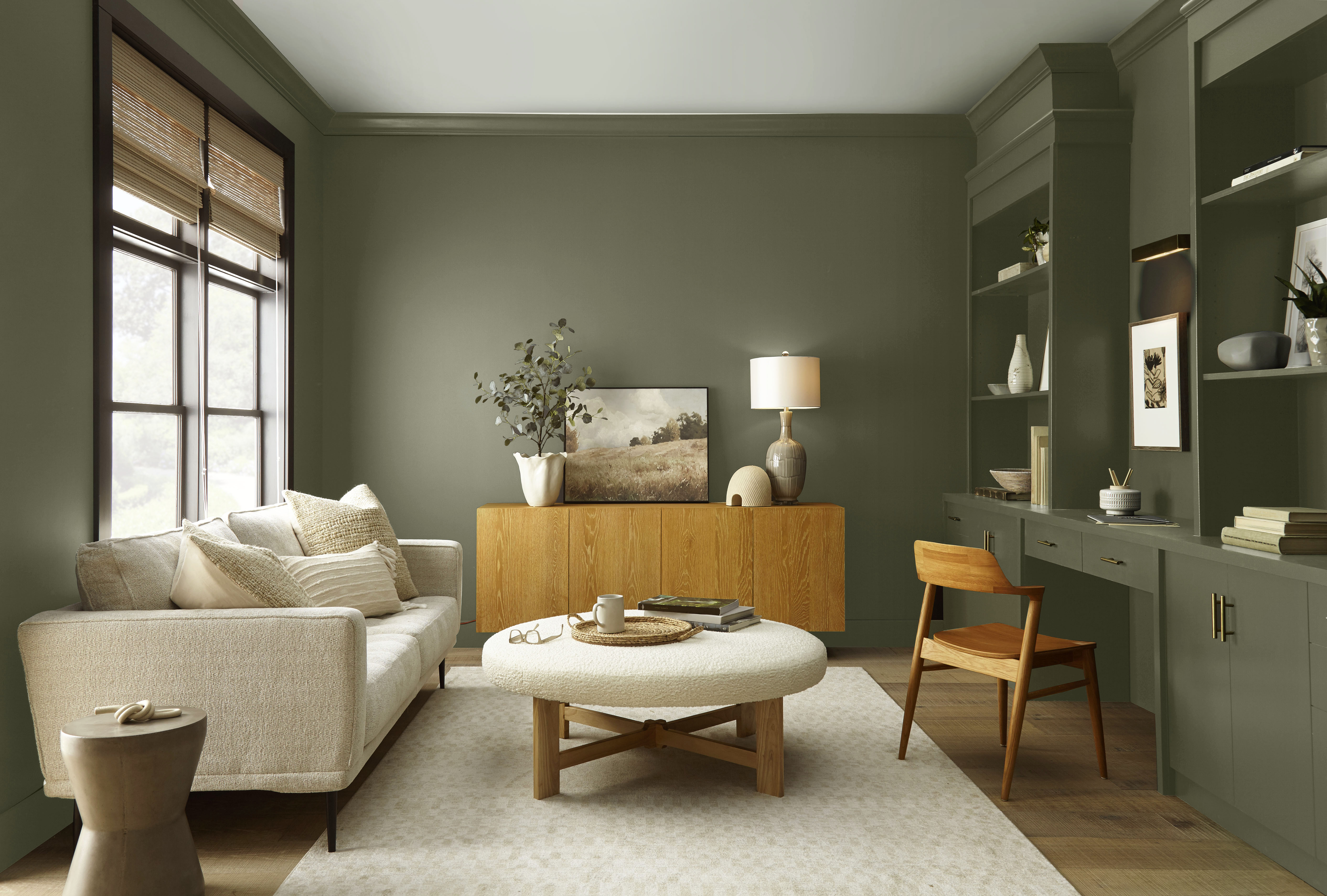 Cette image montre un salon décontracté avec des murs et des étagères encastrées peints dans un vert foncé. Le mobilier comprend un canapé, une table basse, un bahut en bois blond et un coin bureau qui se fond discrètement dans le décor, le tout baigné d’une douce lumière naturelle.