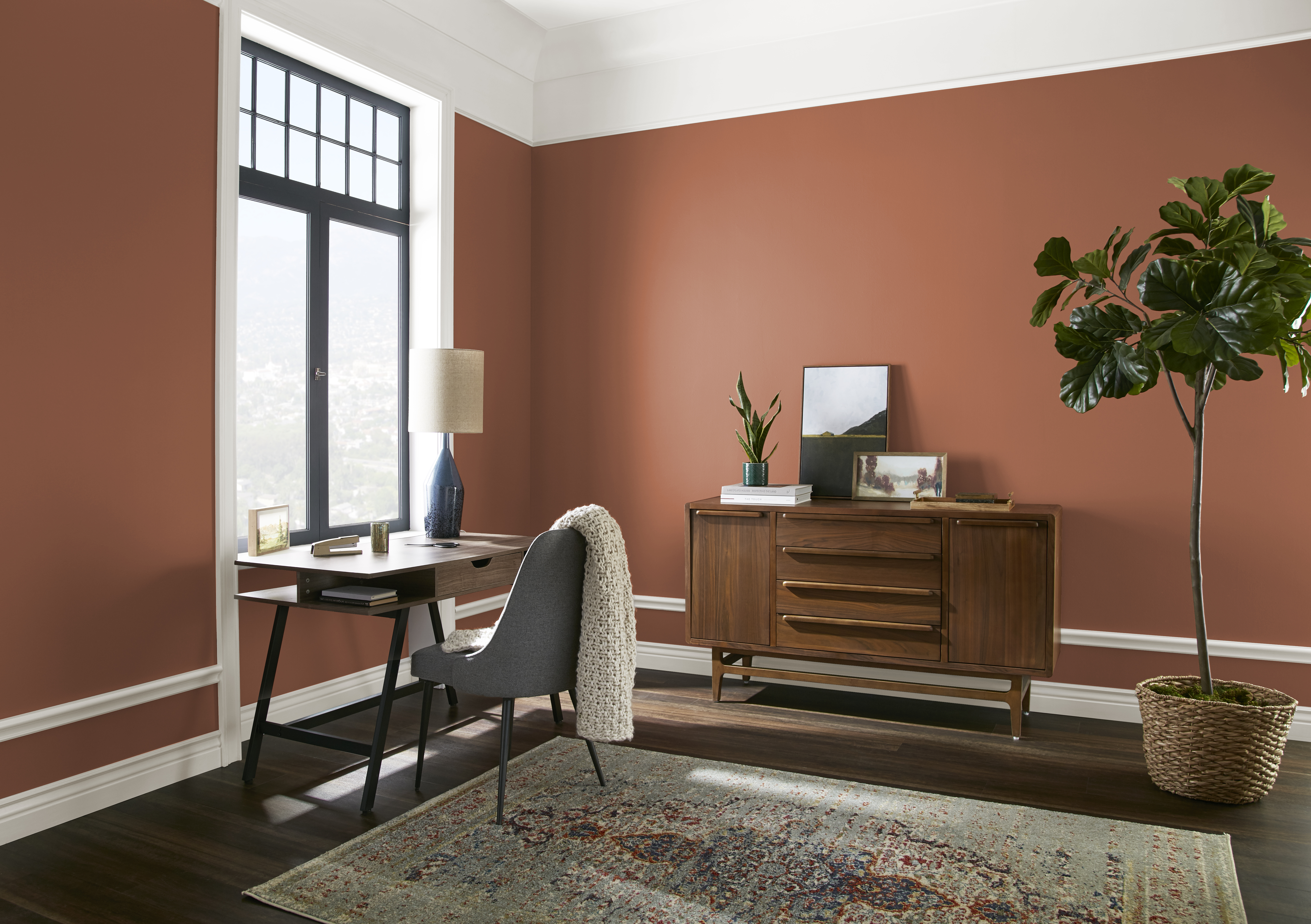 Cette image montre un petit espace de télétravail attenant à une chambre à coucher. Sur fond de murs peints dans une teinte de brique orangée, on aperçoit une table de bureau devant une grande fenêtre, un bahut en bois, un tapis à motifs et une grande plante d’intérieur.