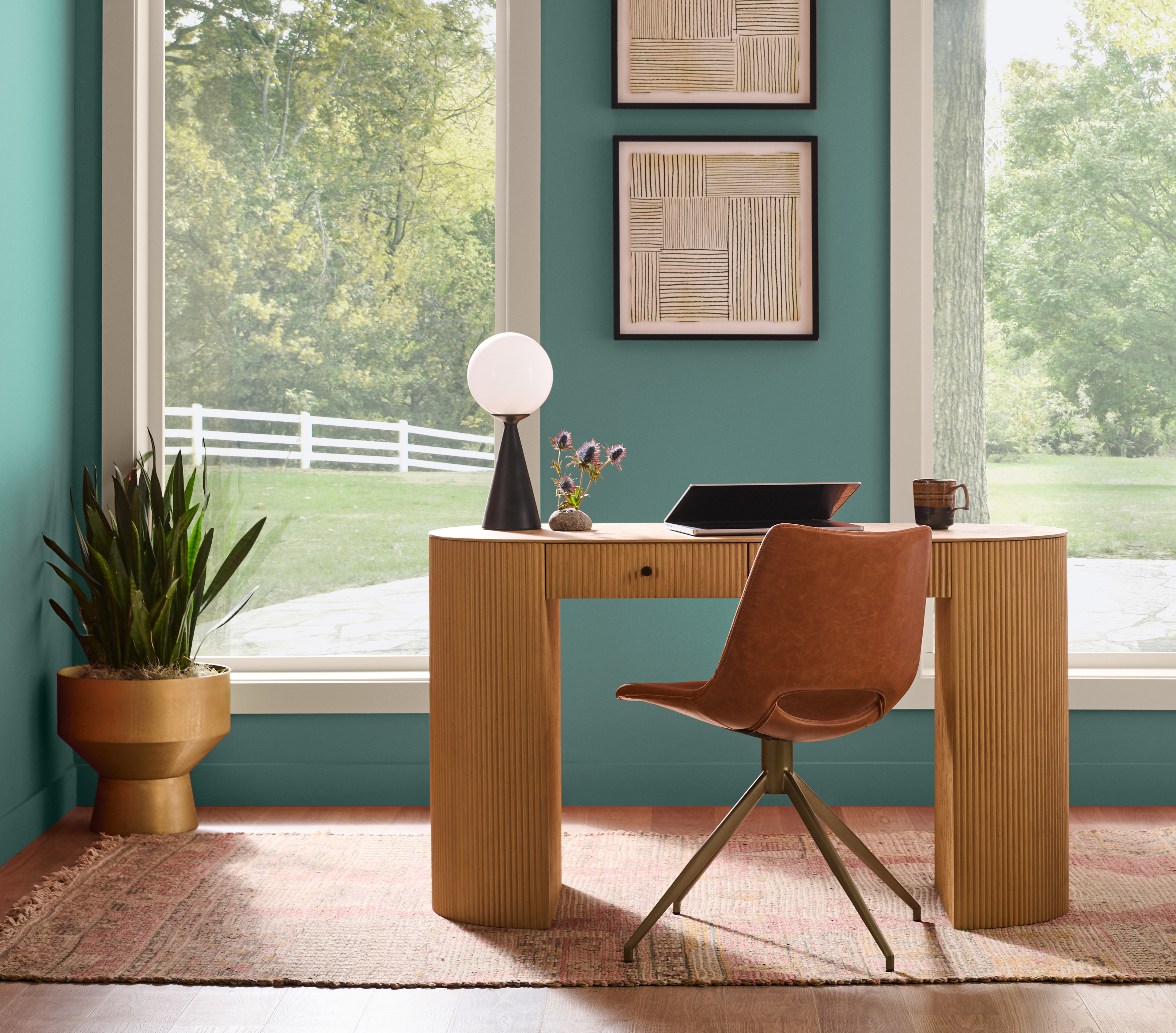 Cette image montre un coin bureau aménagé dans une pièce de séjour à aire ouverte, avec des murs peints en un jade fumé, une petite table de bureau en bois, une chaise moderne, deux immenses fenêtres, des œuvres d’art encadrées et quelques plantes d’intérieur.