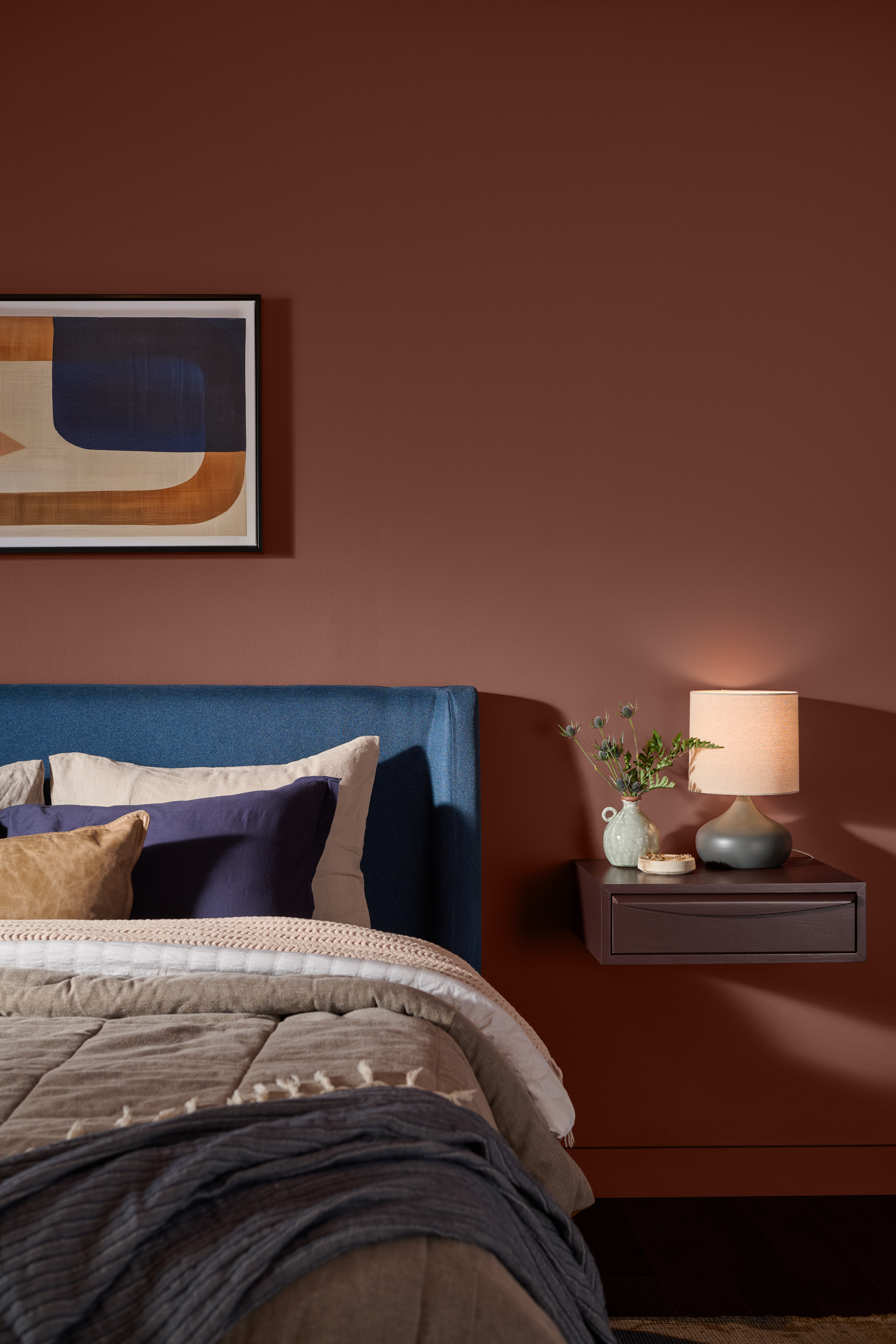 Cette image montre une chambre à coucher avec des murs peints en Terre de Toscane (T27-02), une teinte chaude aux nuances argileuses que l’on a joliment agencé à une literie bleue, grise et beige, à du bois naturel et à un éclairage tamisé.