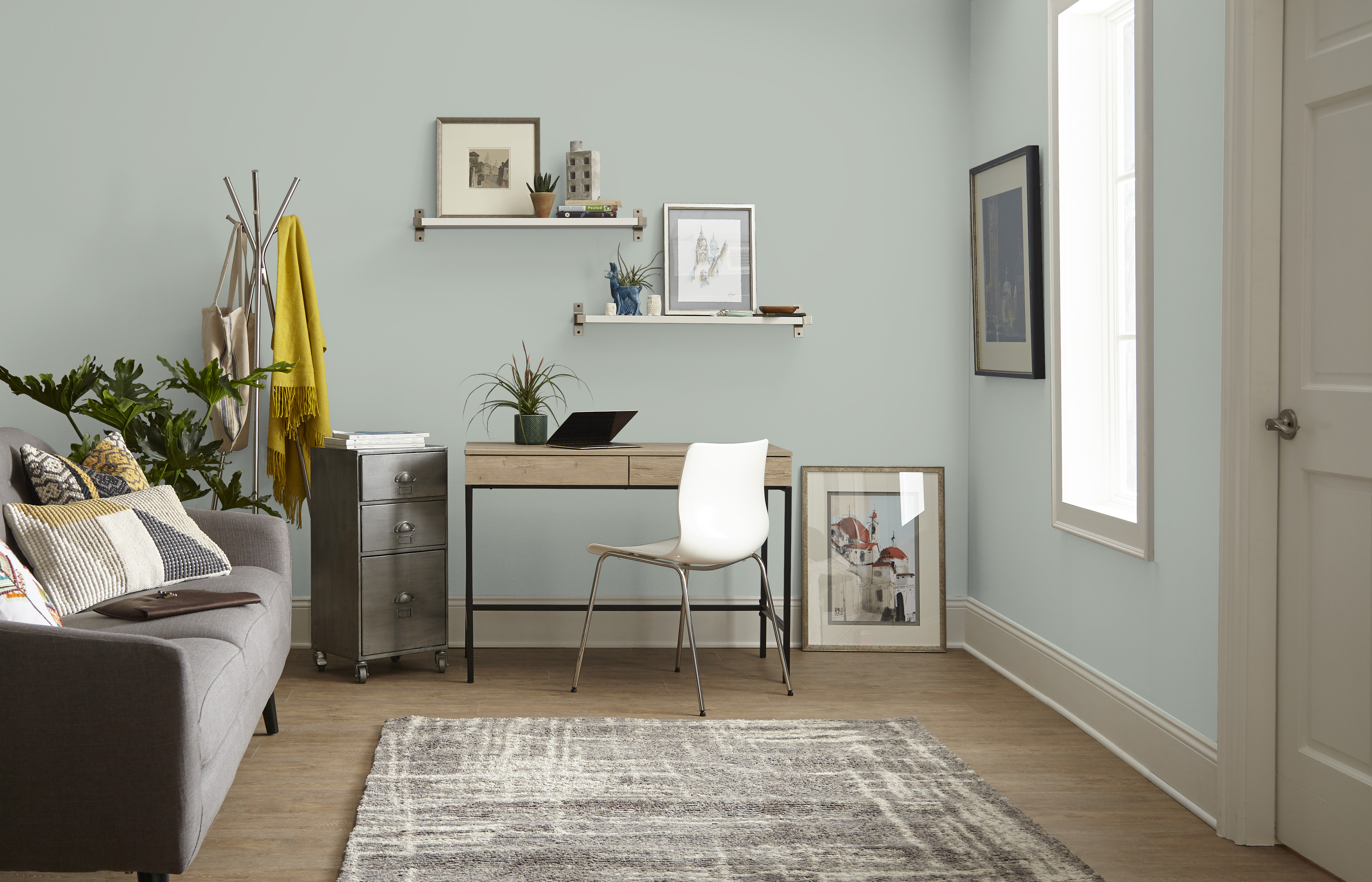 Cette image montre un coin bureau discret aménagé dans un petit salon aux murs peints en gris-vert, une table de bureau compacte, une chaise blanche, deux tablettes flottantes, un canapé, quelques plantes d’intérieur et un parquet en bois clair.