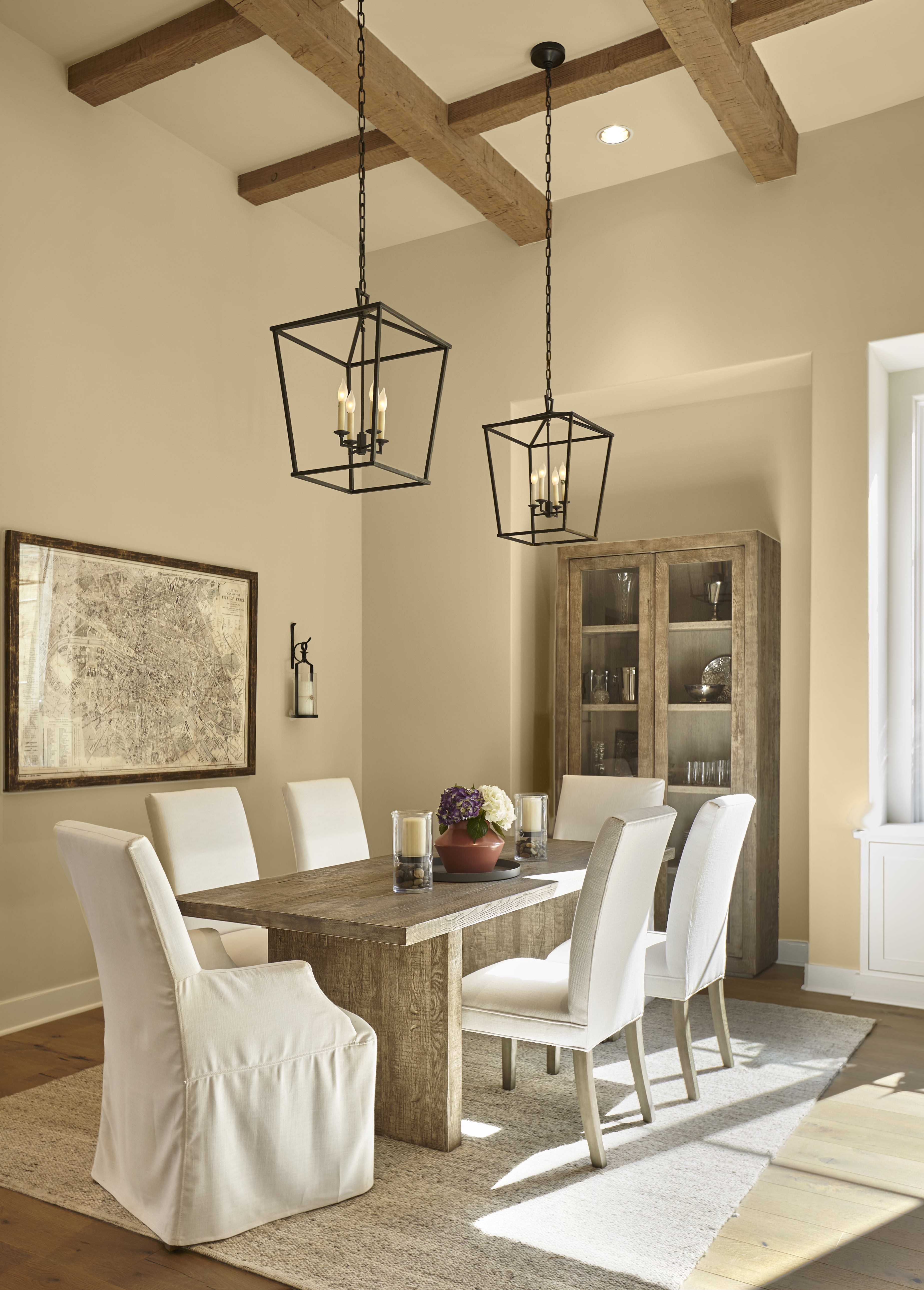 Cette image montre une salle à manger avec des murs peints en Grenier à foin (MQ2-22), une teinte pâle qui rappelle la paille dorée. La pièce est parée d’une table en bois rustique, de poutres apparentes, de chaises de couleur neutre et de luminaires noirs.