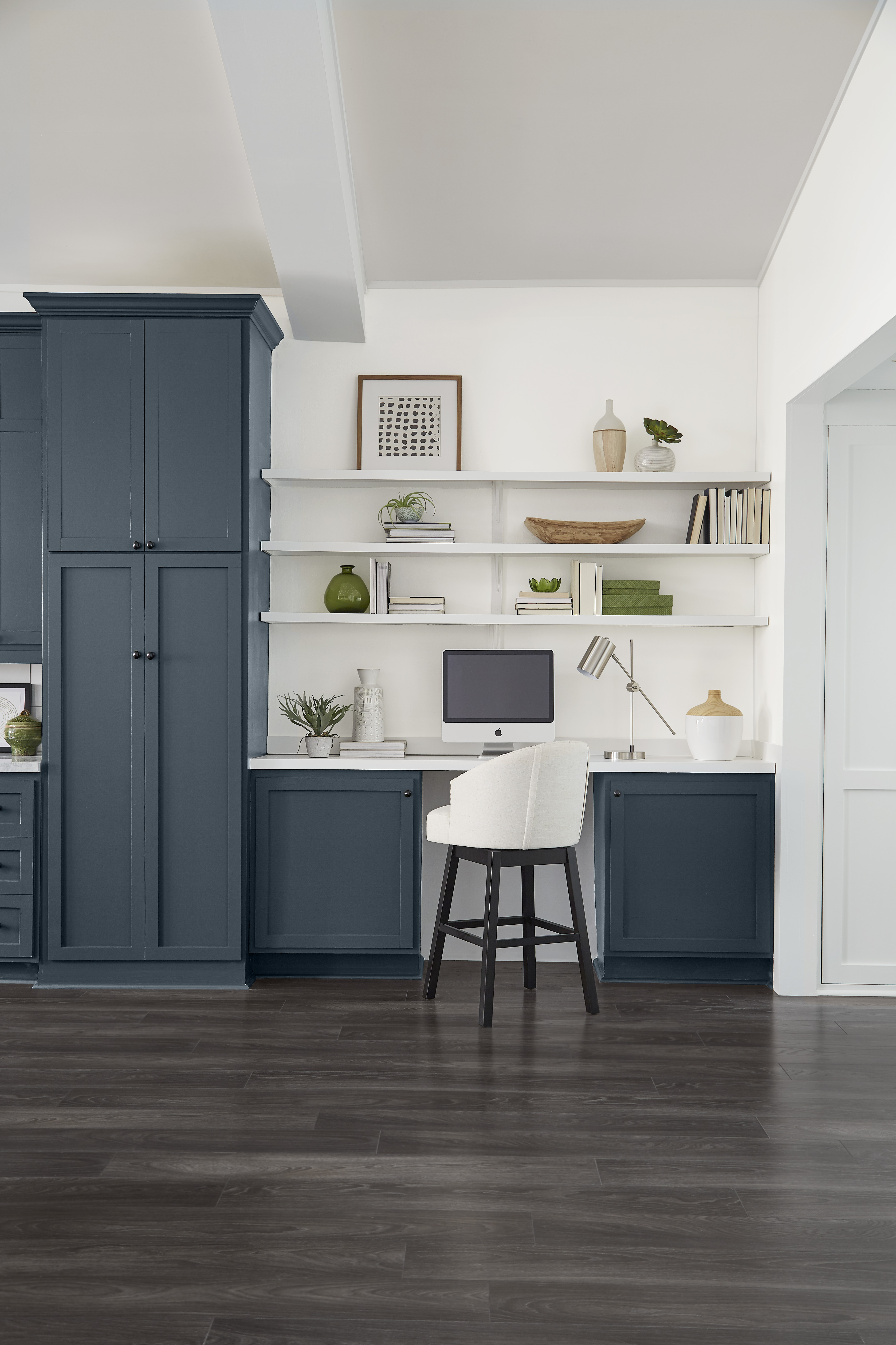 Cette image montre un espace de travail attenant à la cuisine, avec des armoires d’un bleu-gris foncé, une table de travail encastrée, des étagères blanches, une chaise blanche, un écran d’ordinateur et un parquet en bois de couleur foncée.