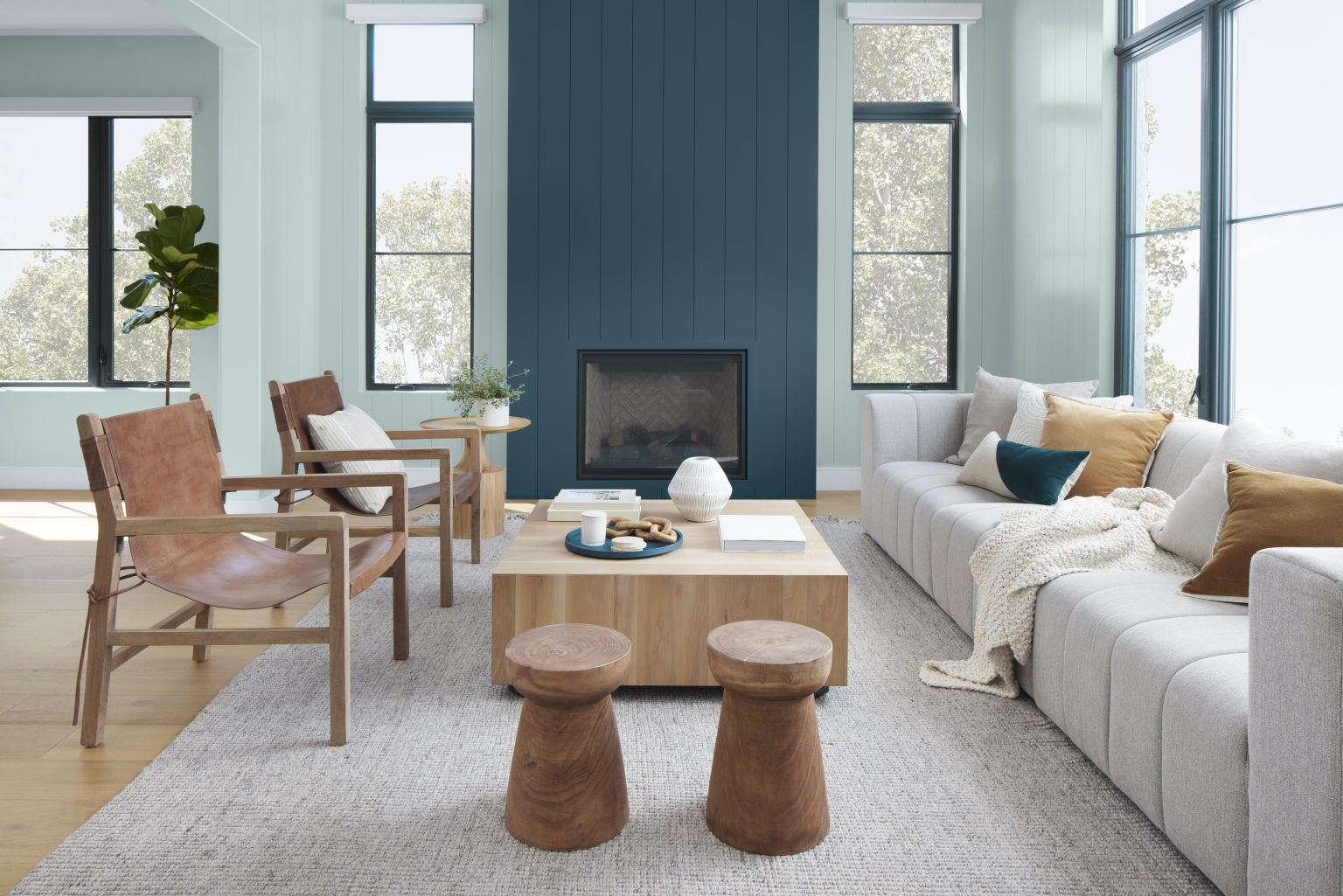 Cette image montre un salon lumineux et contemporain avec des murs peints en teinte Aqueux, un bleu-vert au ton pastel, ainsi qu’une cheminée à panneaux verticaux peints en Bleu nocturne, un bleu profond et sobre. La pièce comporte plusieurs grandes fenêtres aux cadres noirs, des meubles en bois naturel, deux fauteuils en cuir à dossier suspendu, un immense canapé de couleur neutre où sont déposés plusieurs coussins et un jeté, le tout formant un décor aéré, décontracté et empreint d’une ambiance moderne à l’allure côtière rafraîchissante.