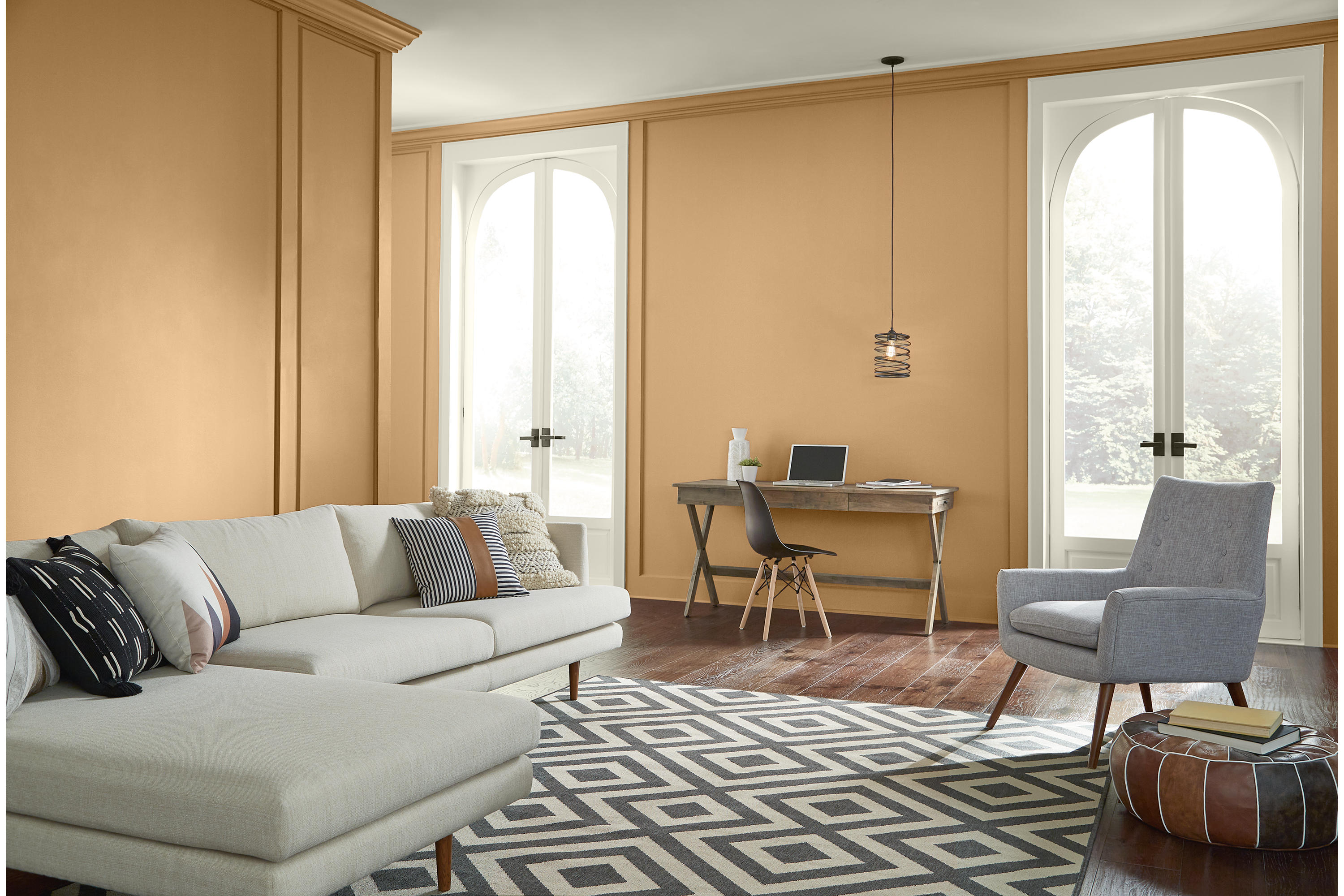 Cette image montre un salon lumineux dont les murs sont peints en teinte Or Cellini, et les boiseries en teinte Café suisse. La pièce comporte un canapé sectionnel de couleur neutre, un fauteuil gris pâle, un plancher de couleur acajou, un tapis à motif géométrique noir et blanc ainsi que deux grandes fenêtres cintrées qui inondent la pièce d’une douce lumière naturelle.