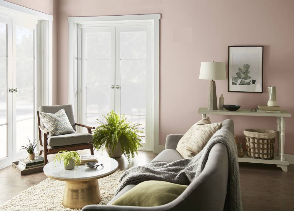 Cette image montre un salon dont les murs sont peints en Rose fumé de BEHRMD avec des moulures peintes en BLANC ULTRA PURMC. Deux grandes portes-fenêtres déversent une abondante lumière naturelle dans la pièce, tandis que le mobilier aux couleurs neutres, les finis en bois au ton chaud et les plantes vertes confèrent à l’ensemble une atmosphère douce et aérée..