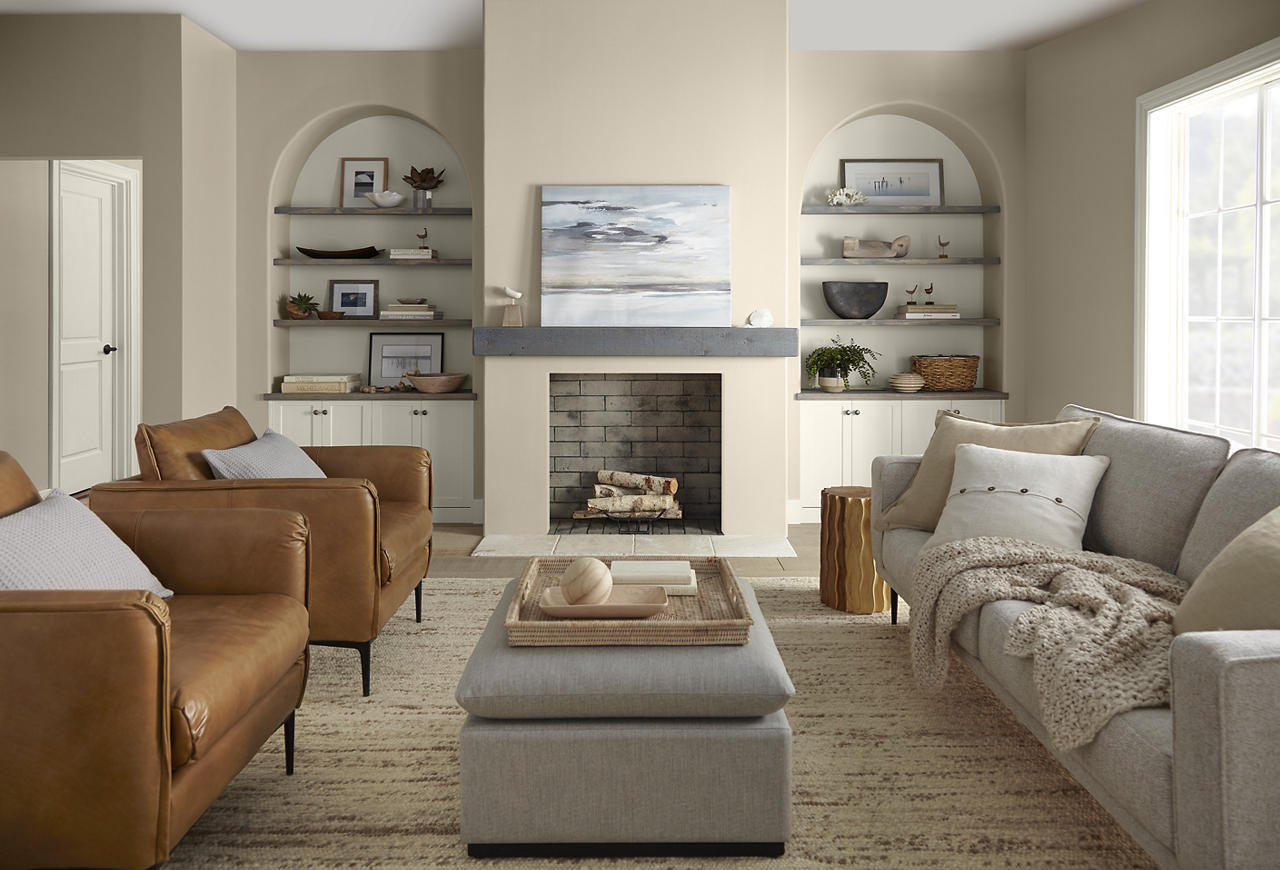 Cette image montre un salon chaleureusement décoré dans le style transitionnel, avec des murs peints en teinte Encore plus beige et des garnitures peintes en Toile vierge. Les fauteuils en cuir de couleur chamois, le canapé gris clair, les étagères encastrées dans des embrasures voûtées, les objets tissés aux tons neutres et le manteau de cheminée classique créent un décor confortable et harmonieux. La lumière naturelle abondante, jumelée à la palette de beiges et de blanc crème, confère à la pièce une ambiance accueillante, détendue et empreinte d’une sophistication subtilement moderne.