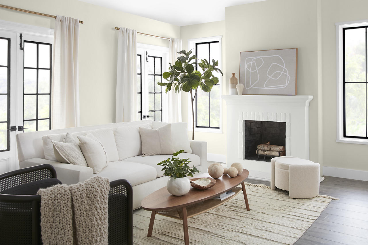 Cette image montre un salon lumineux au décor épuré, avec des murs peints en teinte Toile vierge, un blanc crème doux aux nuances chaudes et accueillantes qui crée une impression d’équilibre, sans paraître jaunâtre ni austère. La teinte BLANC ULTRA PURMC met en valeur les garnitures et la cheminée, tandis que les fenêtres à croisillons noirs, le canapé et le pouf blancs, les accents de bois clair et la lumière naturelle abondante viennent parachever l’ensemble.