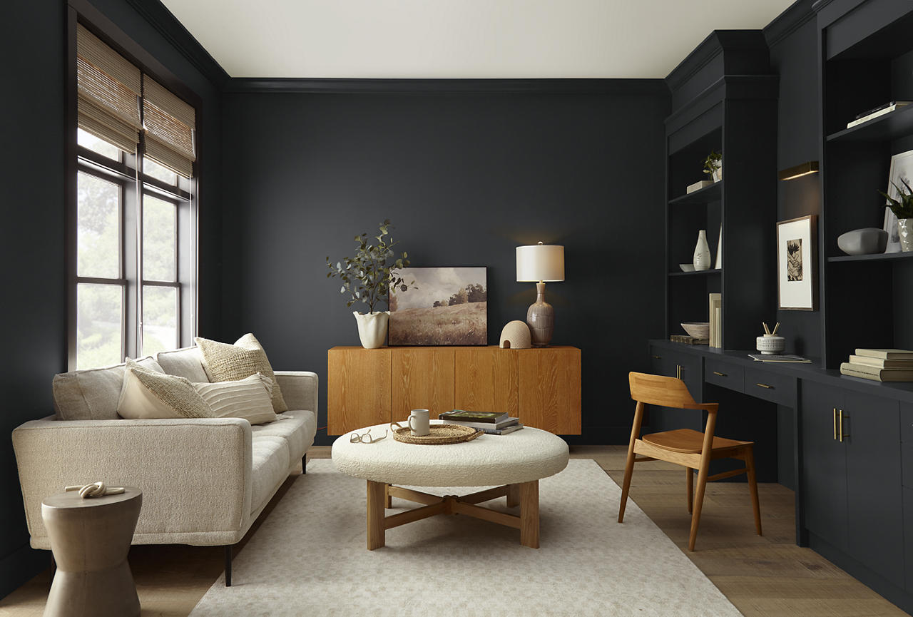 Cette image montre un salon moderne à l’ambiance intimiste, dont les murs, les garnitures, le bureau encastré et les armoires sont peints en teinte Poivre concassé, un noir doux qui crée une toile de fond riche et enveloppante. Le canapé de couleur neutre, les meubles en bois blond, les matières textiles également de couleur neutre et la lumière naturelle entrant par les deux grandes fenêtres viennent adoucir la palette de teintes foncées. Ici, les éléments encastrés et le mobilier aux lignes épurées confèrent à l’espace une ambiance raffinée et contemporaine.