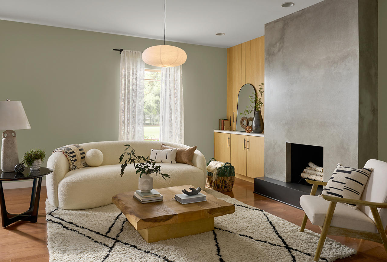 Cette image montre un salon de style contemporain dont les murs sont peints en teinte Camouflage de jungle, et le plafond en teinte Blanc soyeux. On aperçoit une cheminée en plâtre à l’aspect moderne, des armoires basses encastrées en bois blond, un canapé en demi-lune au ton neutre, des objets décoratifs aux textures naturelles et une abondante lumière naturelle.