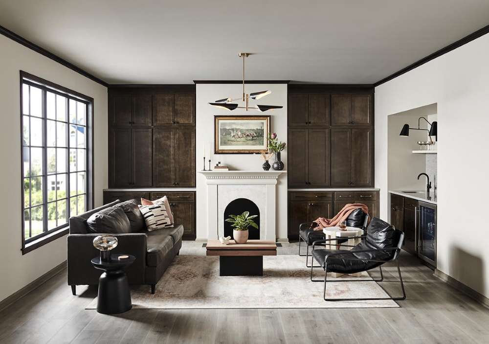Cette image montre un salon contemporain au style raffiné, avec des murs peints en teinte Gris tranquille et des garnitures peintes en NOIR. La pièce comporte une immense fenêtre à croisillons noirs et une cheminée blanche flanquée d’armoires encastrées en bois foncé. L’ameublement consiste en un divan et deux fauteuils en cuir noir avec des objets décoratifs en bois au ton chaud, le tout formant un style soigné et hautement contrasté. La douce lumière naturelle et le jeu des teintes neutres et sobres confèrent à l’espace une ambiance équilibrée, invitante et toute en finesse.