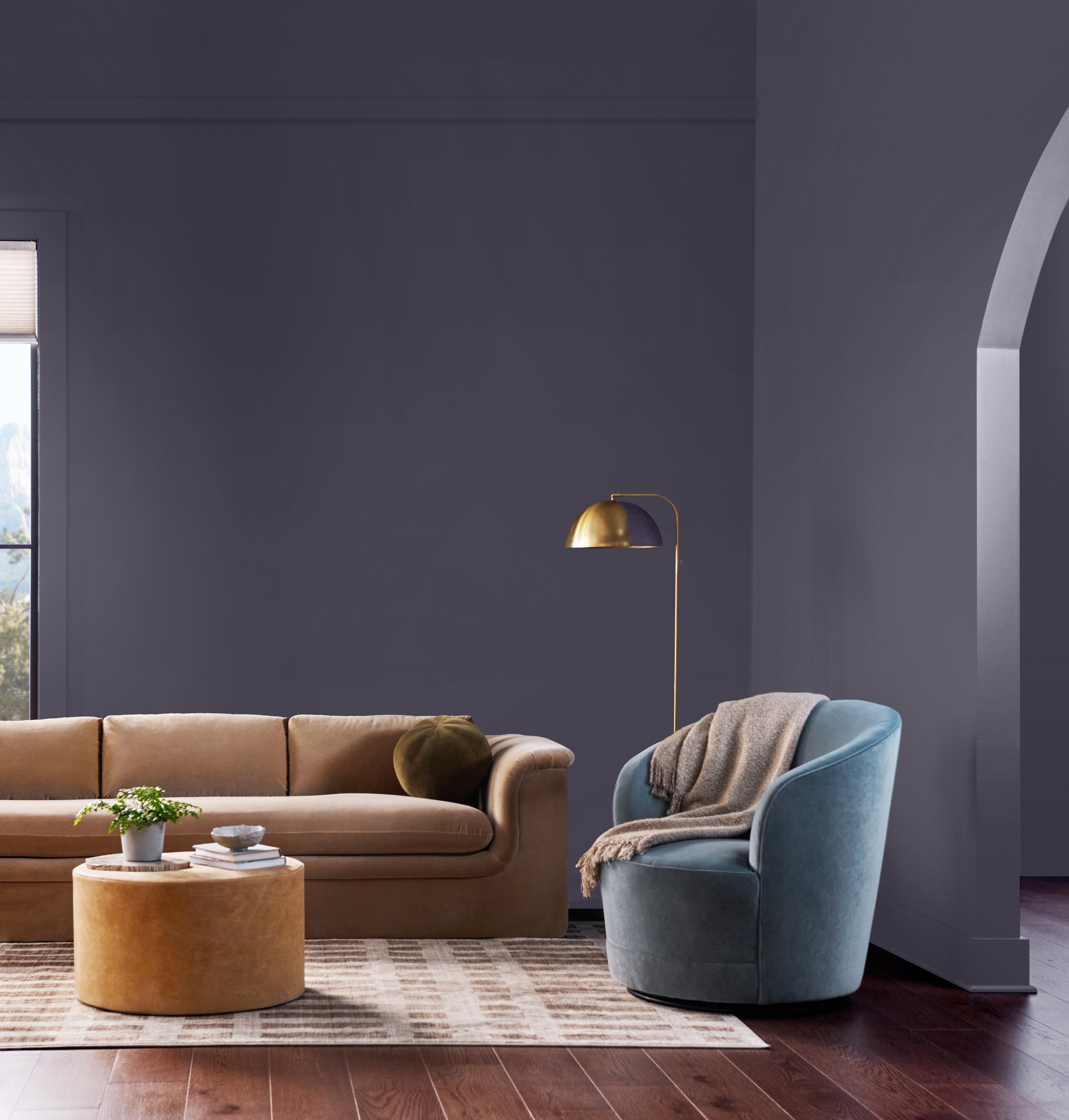 Cette image montre un salon contemporain aux murs peints en Violet opulent, une riche et somptueuse teinte de violet. On y aperçoit un canapé de couleur chamois, un fauteuil d’accent bleu clair, une grande lampe sur pied en laiton, un plancher en bois foncé et une impressionnante embrasure en forme d’arche qui donne sur la pièce voisine.