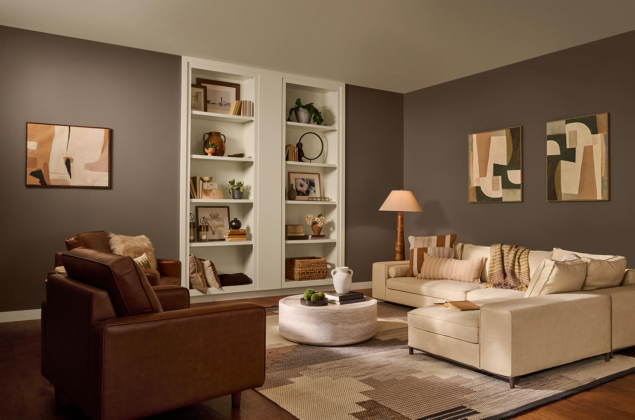 Cette image montre un salon de sous-sol au décor sophistiqué mais non moins chaleureux, dont les murs peints en teinte Introspection contrastent avec le plafond, les moulures et les étagères peints en Beige côtier. La pièce comporte un large canapé modulable de couleur blanc crème, des fauteuils en cuir d’un brun profond, un jeu de coussins et de jetés aux tons neutres, des objets en bois et des œuvres d’art abstraites, qui créent un décor douillet et bien ficelé. Ensemble, la palette de bruns et de beiges, les étagères encastrées et l’éclairage tamisé confèrent à l’espace une ambiance raffinée et intimiste, avec une touche moderne et organique.