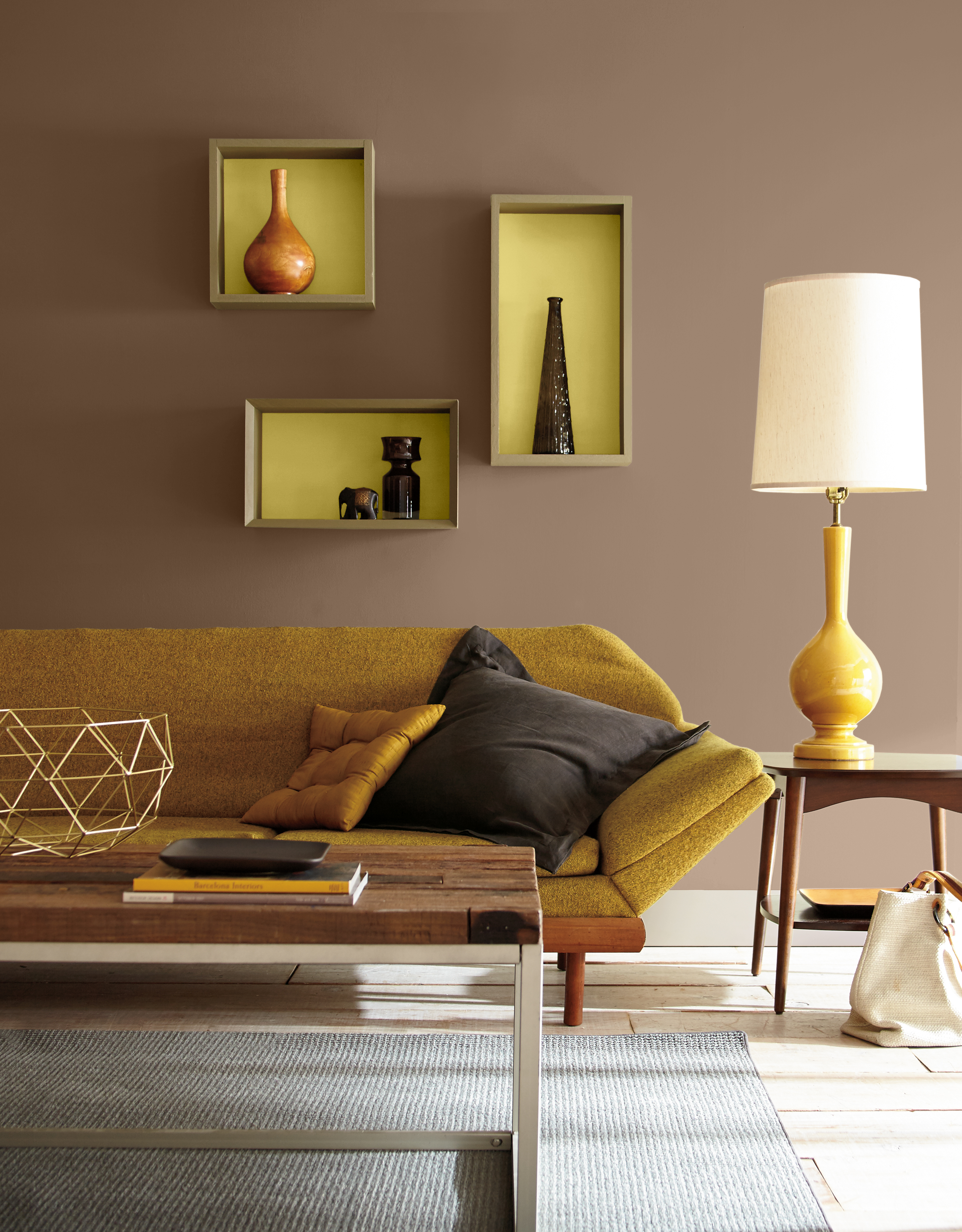 Cette image montre un salon d’inspiration rétro avec des murs peints en Joshua Tree, un brun terreux et chaleureux, agrémenté par la teinte Carambole, un jaune vif tirant légèrement sur le vert que l’on a appliqué à l’intérieur des niches d’exposition encastrées. Un canapé bas de couleur moutarde, une lampe de table de style rétro, une table basse sculpturale en bois et en métal, des objets décoratifs de différentes formes et un mobilier de style milieu du siècle contribuent à créer un décor à l’allure nostalgique et éclectique.