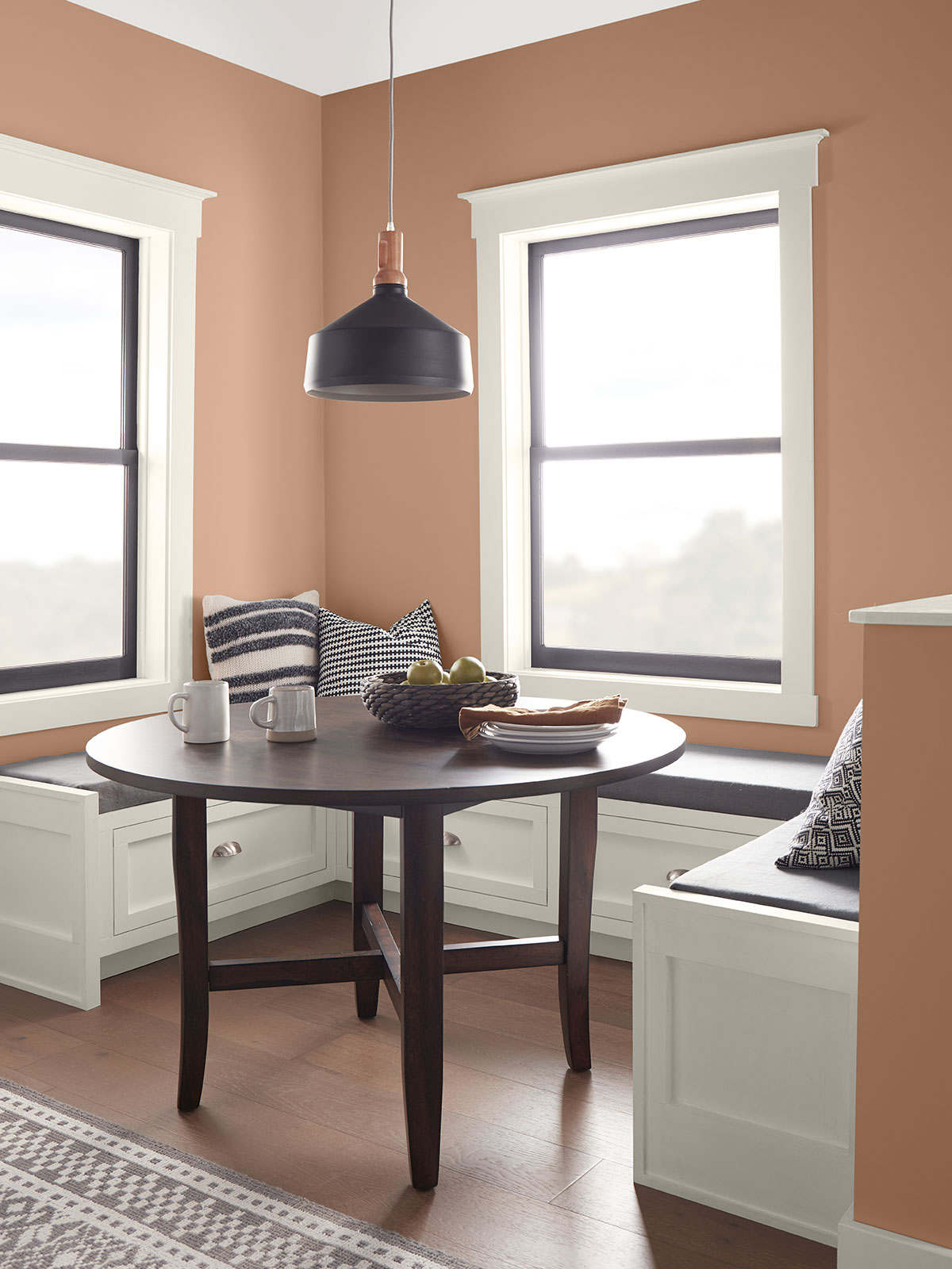A kitchen nook with white built-in seats sitting below two windows. The table is a dark wood tone. The walls are painted in Canyon Dusk.