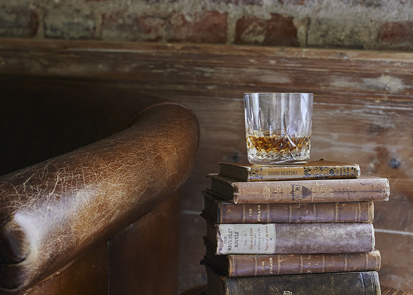 A stack of books with a glass of whisky on top next to a worn leather chair. 