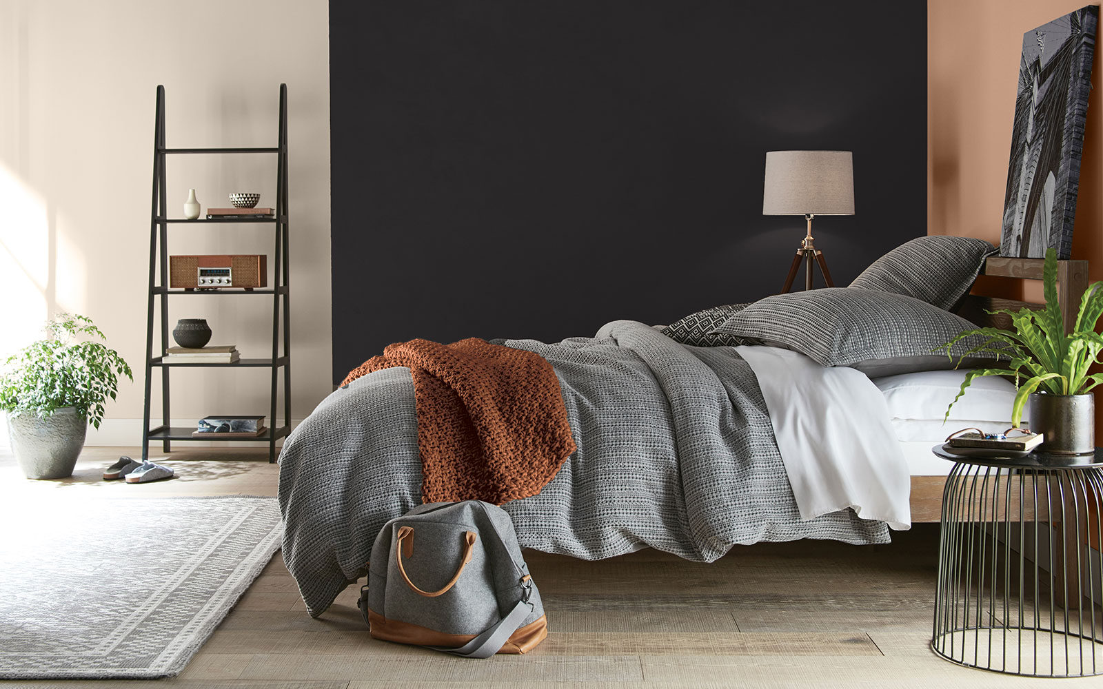 A bed room image with four color swatches sitting beside it. The image displays the four colors: A black hue is used on the side wall of the bedroom, a creamy white hue is used for a wall just outside of the bed area, a terra-cotta orange is used for a blanket, and a dusty orange hue is used on the headboard wall.