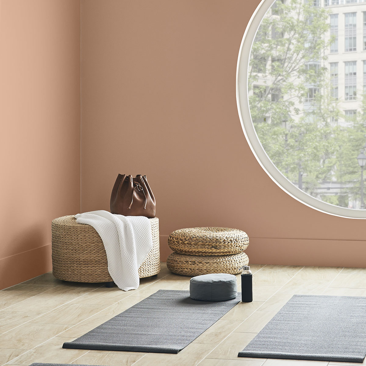 A cropped corner of a workout room with walls painted in Canyon Dusk. There is a large circle window on the wall bringing in a lot of light to the room. A rattan ottoman with a towel and purse placed on top sits in the corner. On the ground are yoga mats.