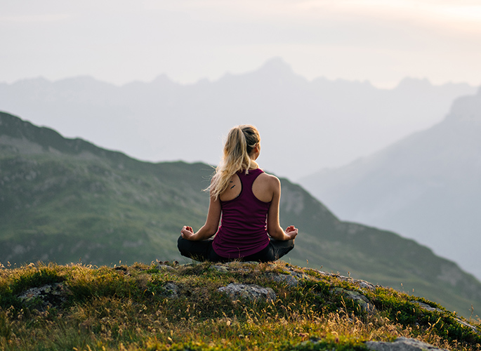 A woman siting on a flowered mountain top meditating. Her surroundings are of a quiet sky and mountains.