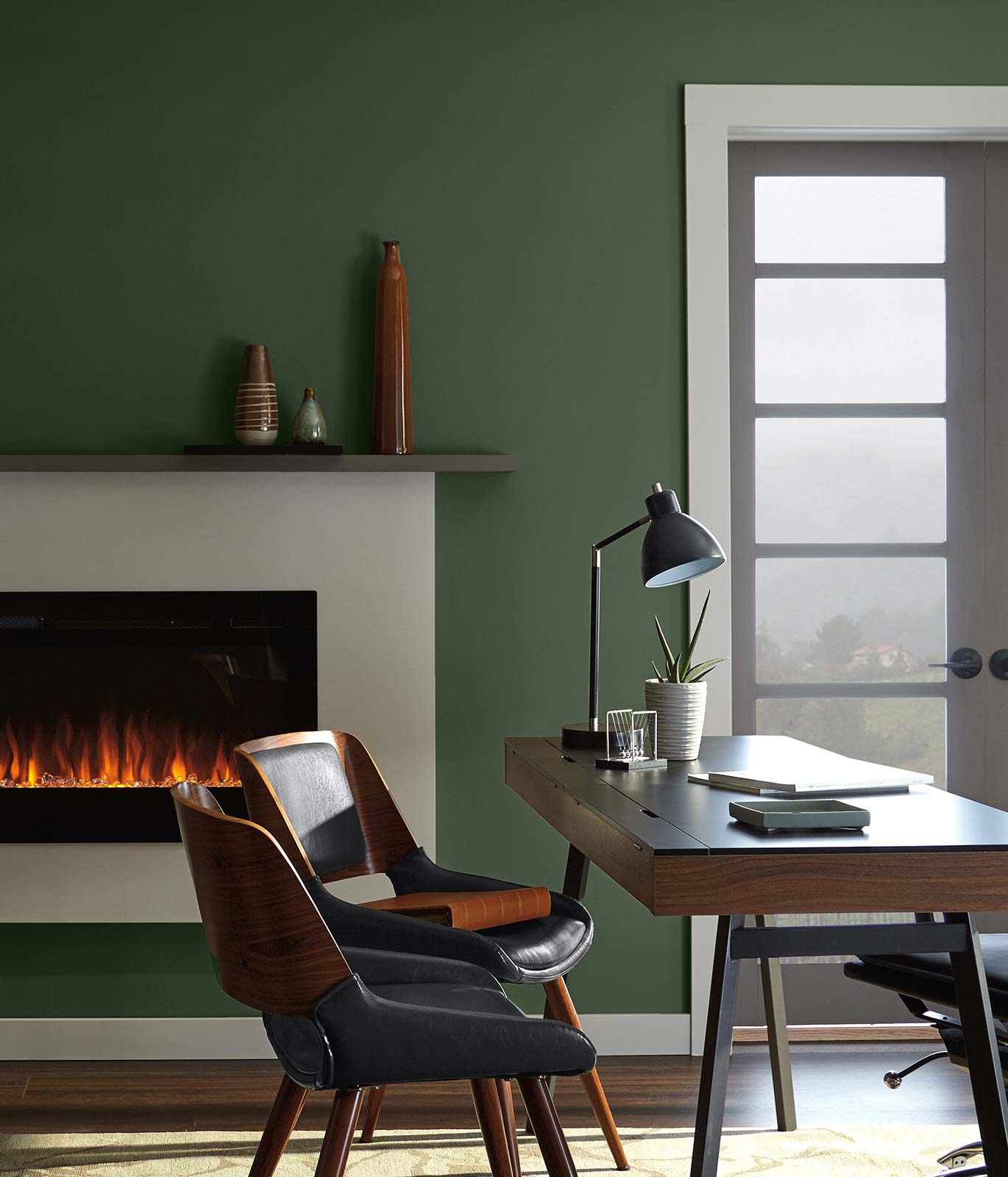 An office desk with a dark brown desk placed in front of a lit fireplace. Walls in the room are painted green. The mood is quiet and focused.
