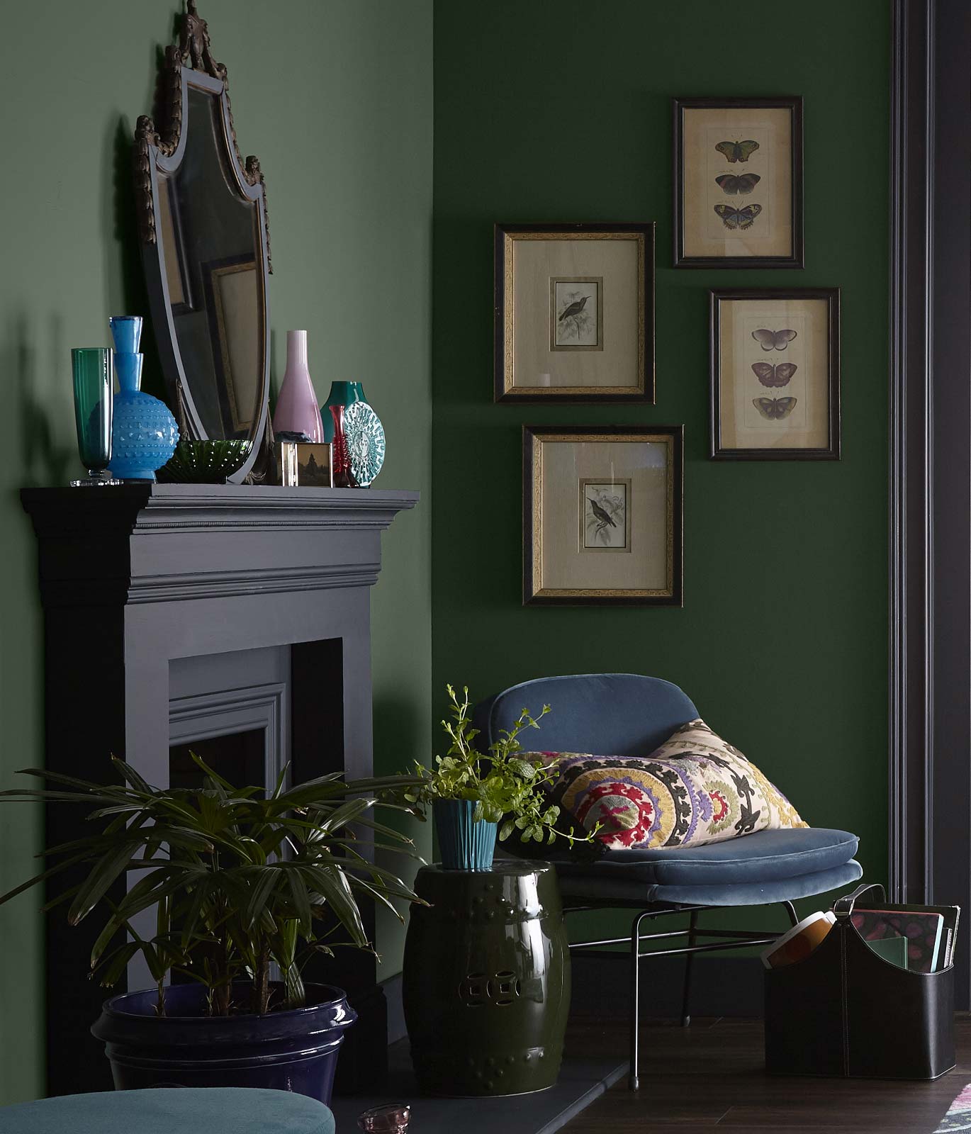 A quiet corner of a living room showing a black painted fireplace, a velvety chair in a blue color. The walls of the room are painted in a dark green color.